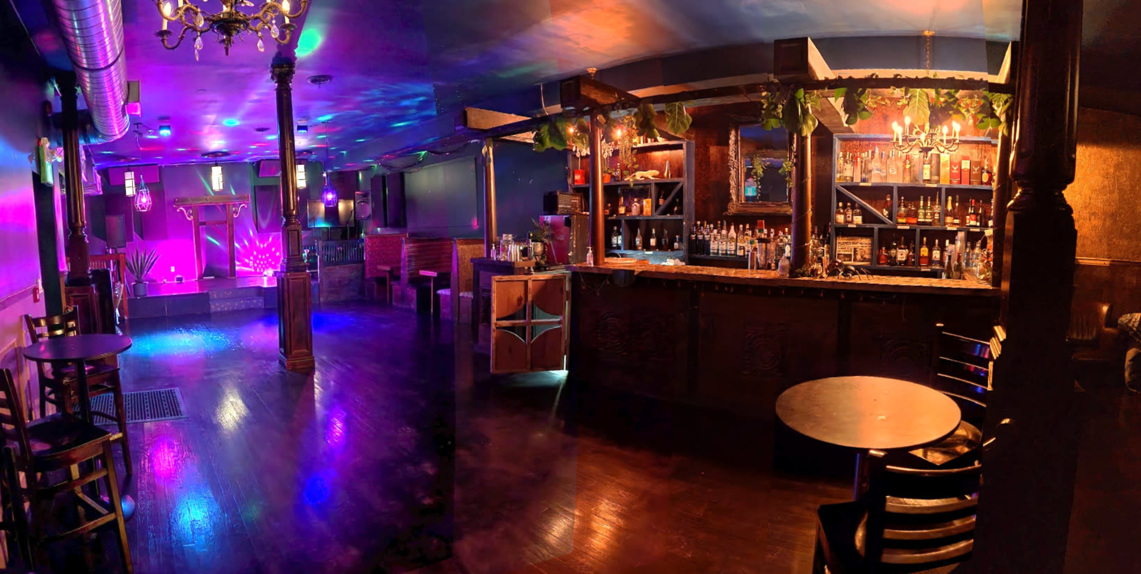 The image shows a dimly lit bar area with a wooden counter, shelves stocked with bottles, and colorful lighting across the floor and walls.