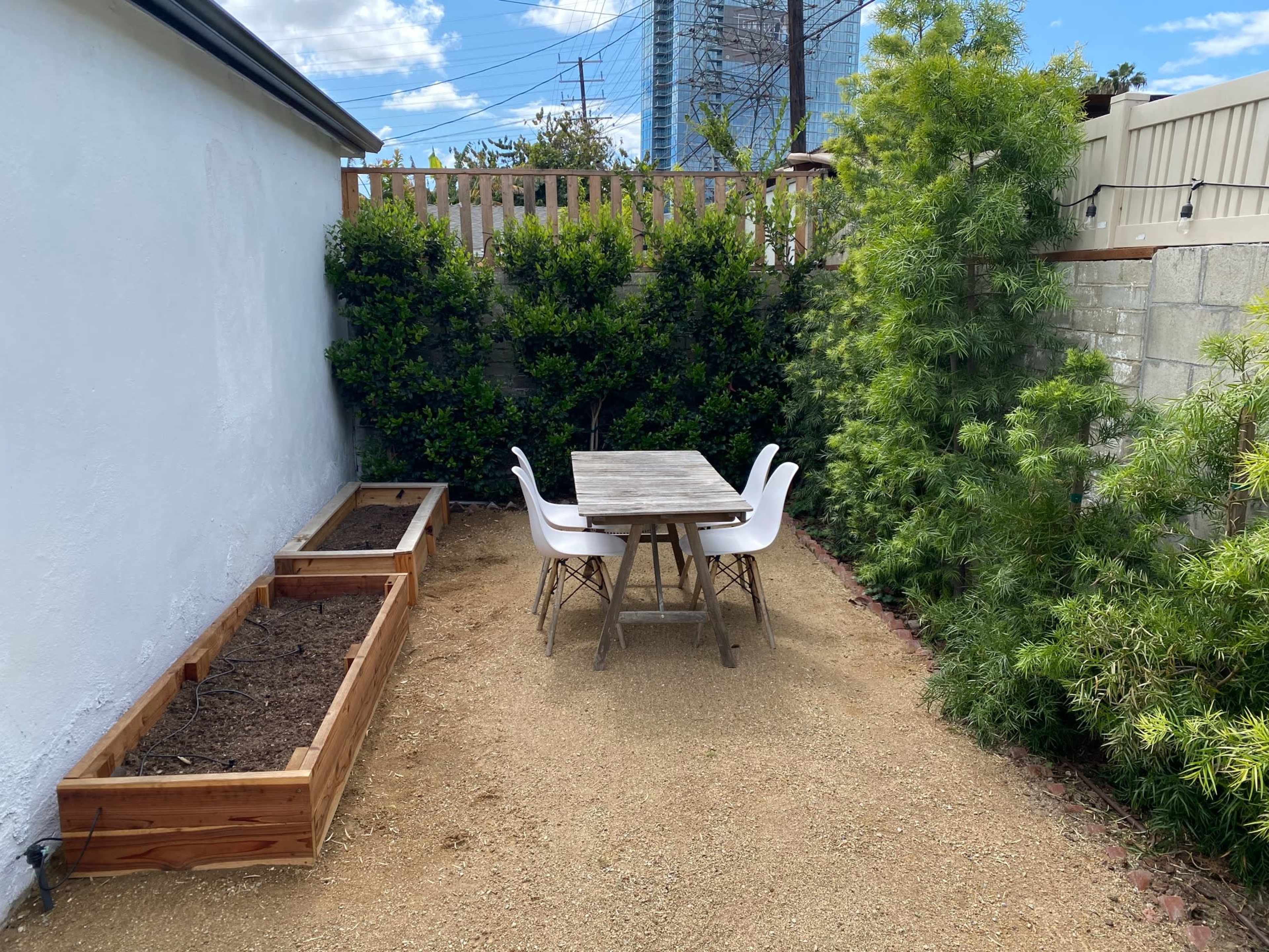 A patio area features a wooden table and chairs surrounded by gravel and two raised garden beds.