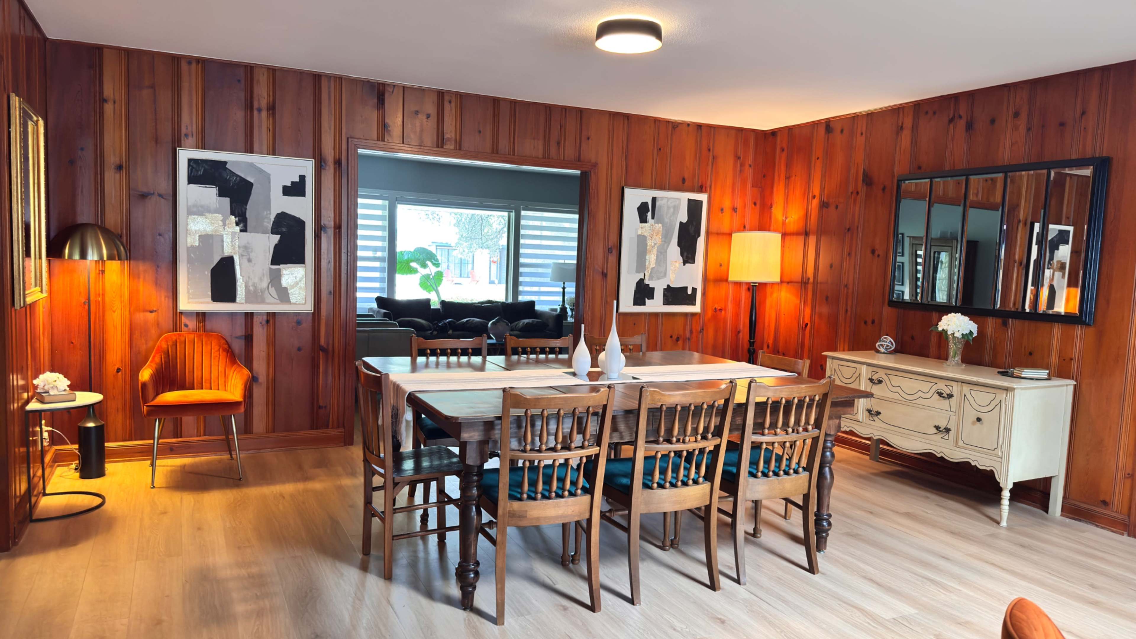 The image shows a dining area with wooden walls, a long table surrounded by chairs, and contemporary art pieces on display.
