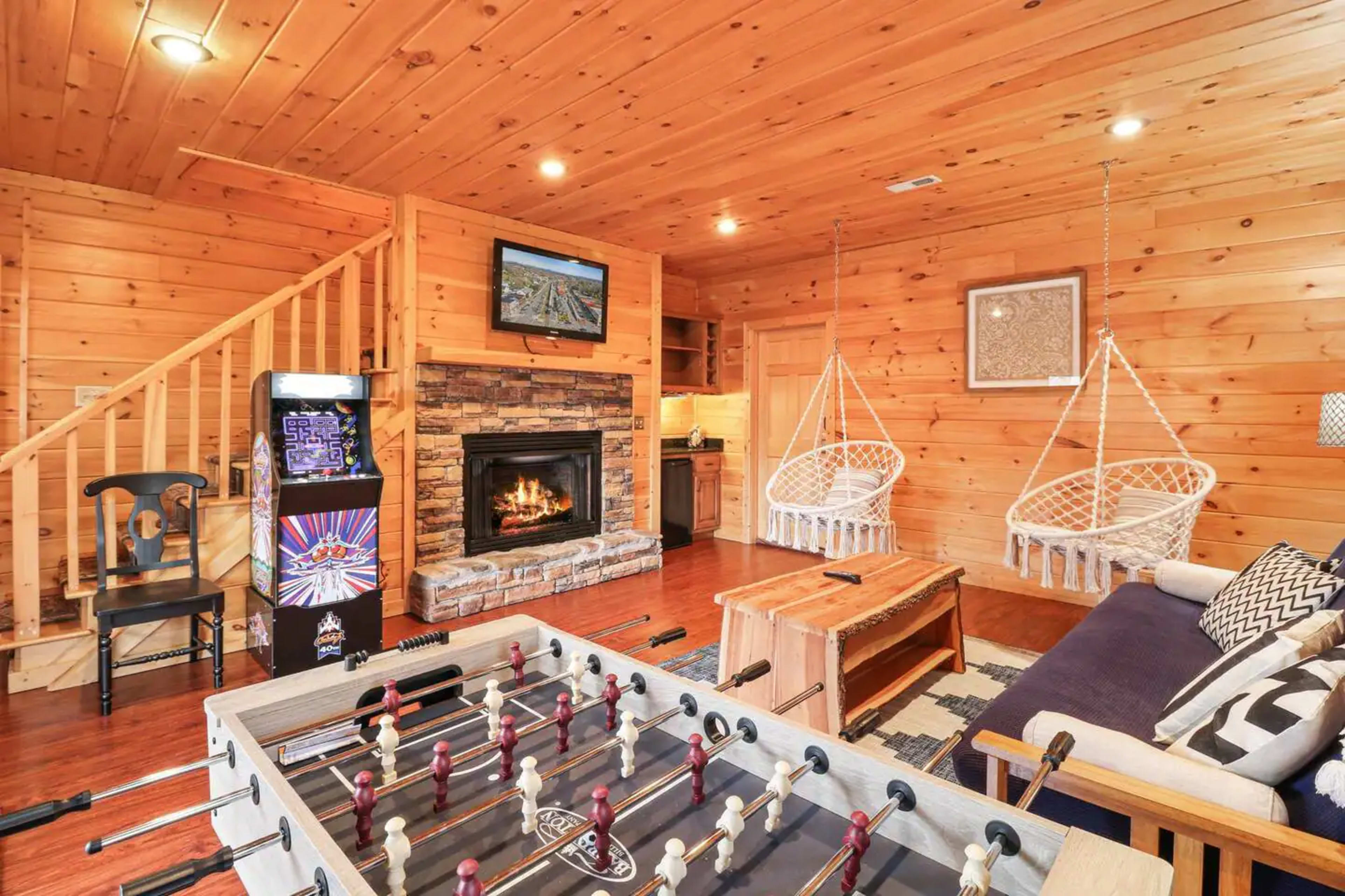 The room features a wooden interior with a foosball table in the foreground, a stone fireplace, two hanging chairs, and stairs leading to an upper level.