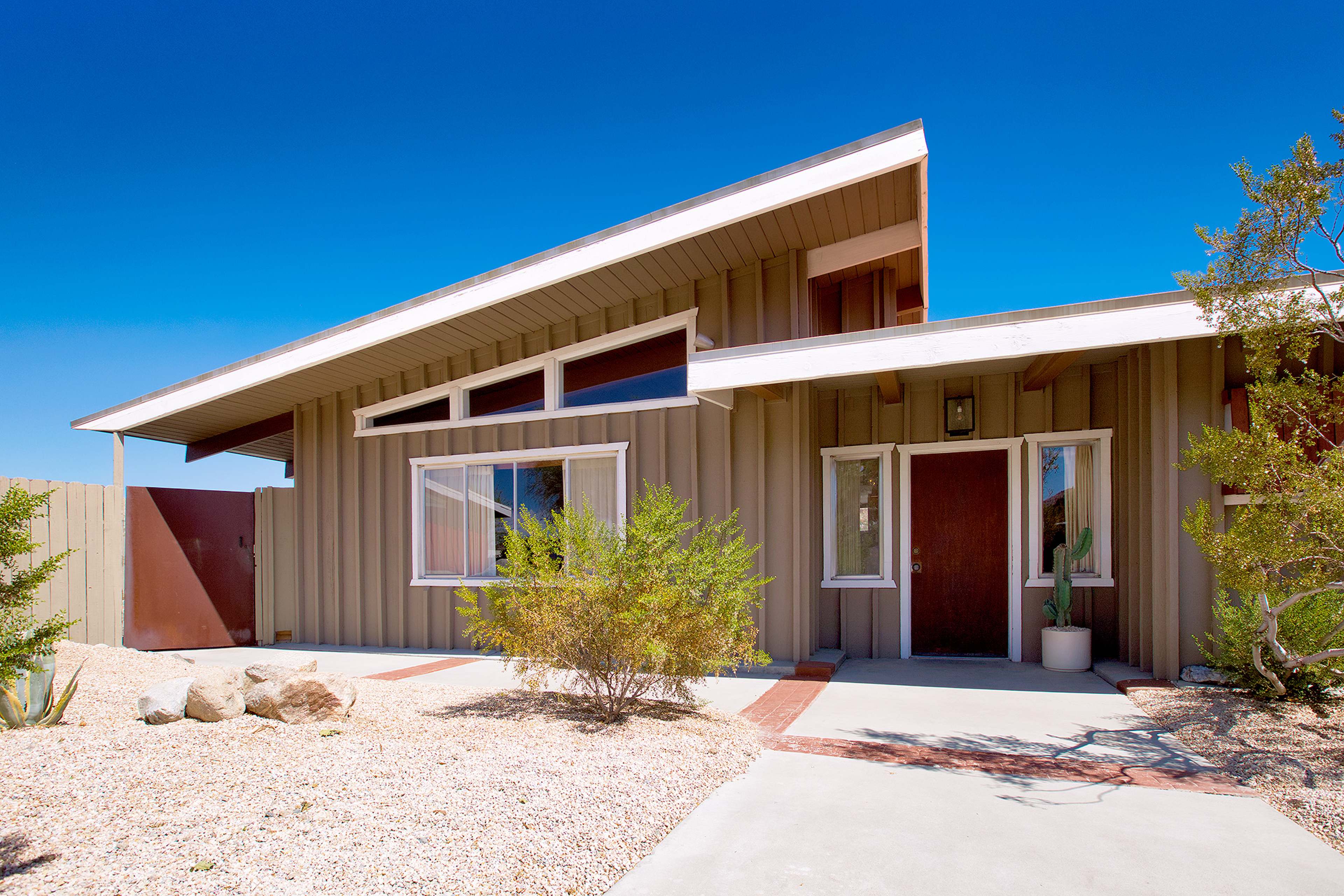 A modern, single-story house with a sloped roof, surrounded by gravel landscaping and low plants against a clear blue sky.