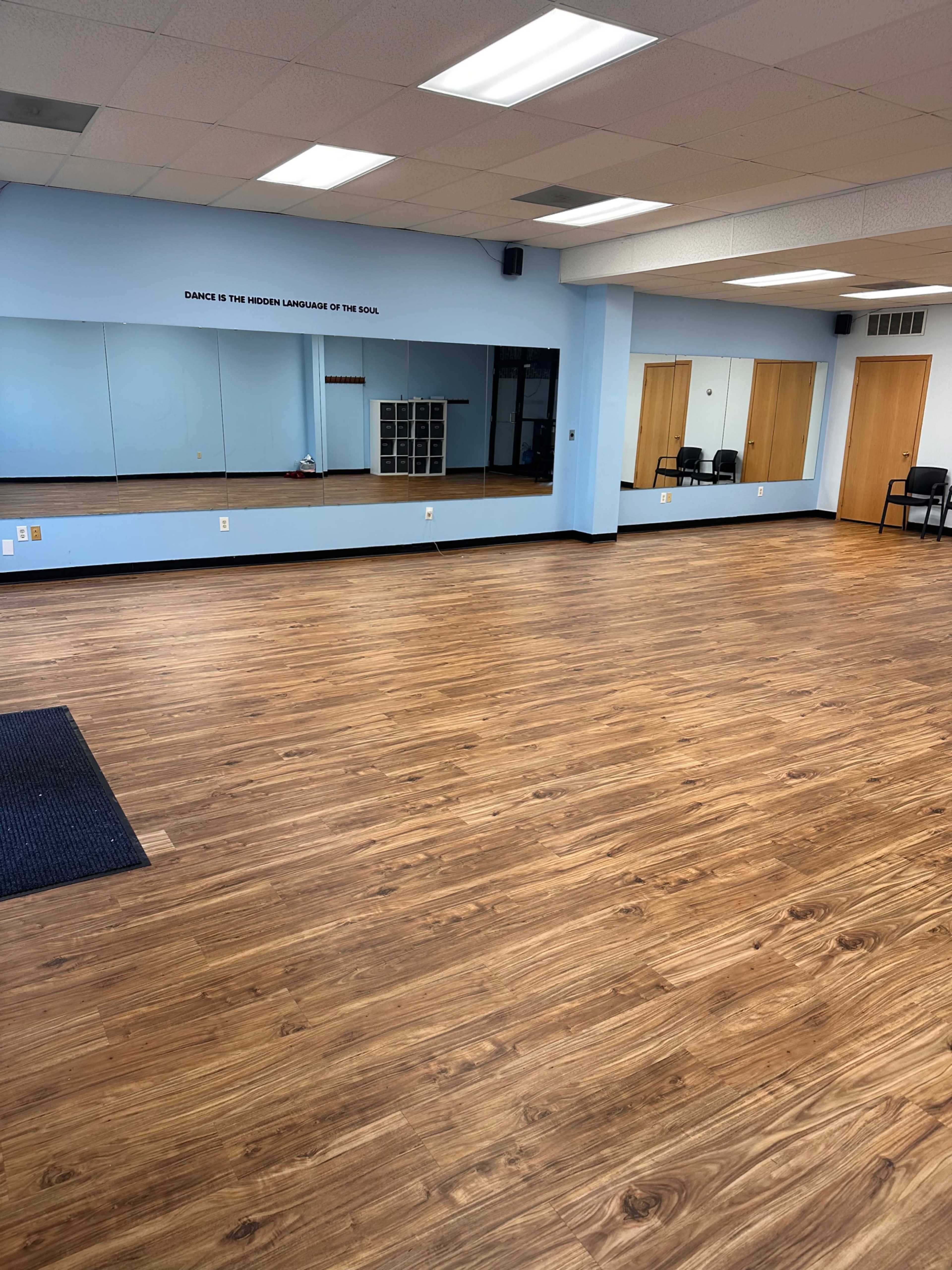 A dance studio with wooden flooring, mirrors on one wall, and a few chairs in the background.