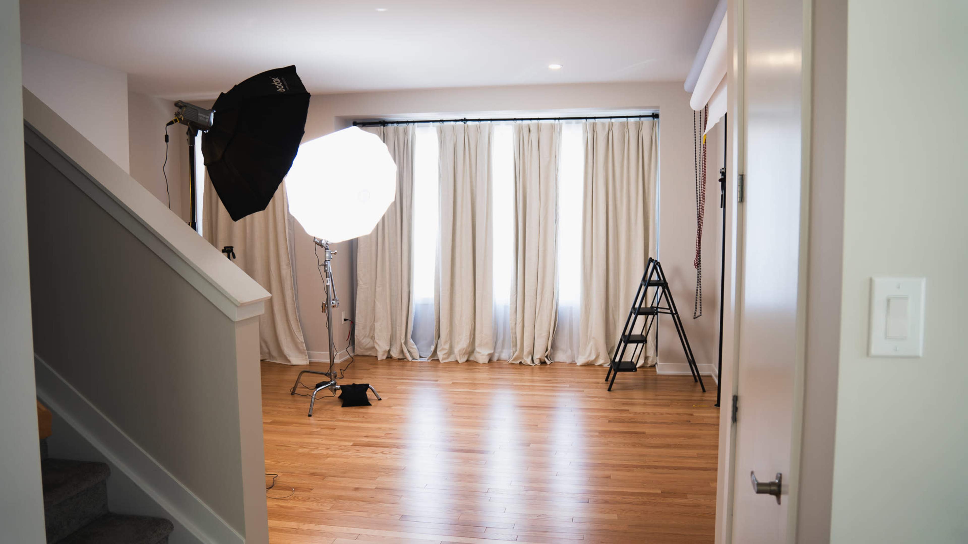 A bright, open room with wooden flooring, large windows covered by white curtains, and photography equipment set up in one corner.