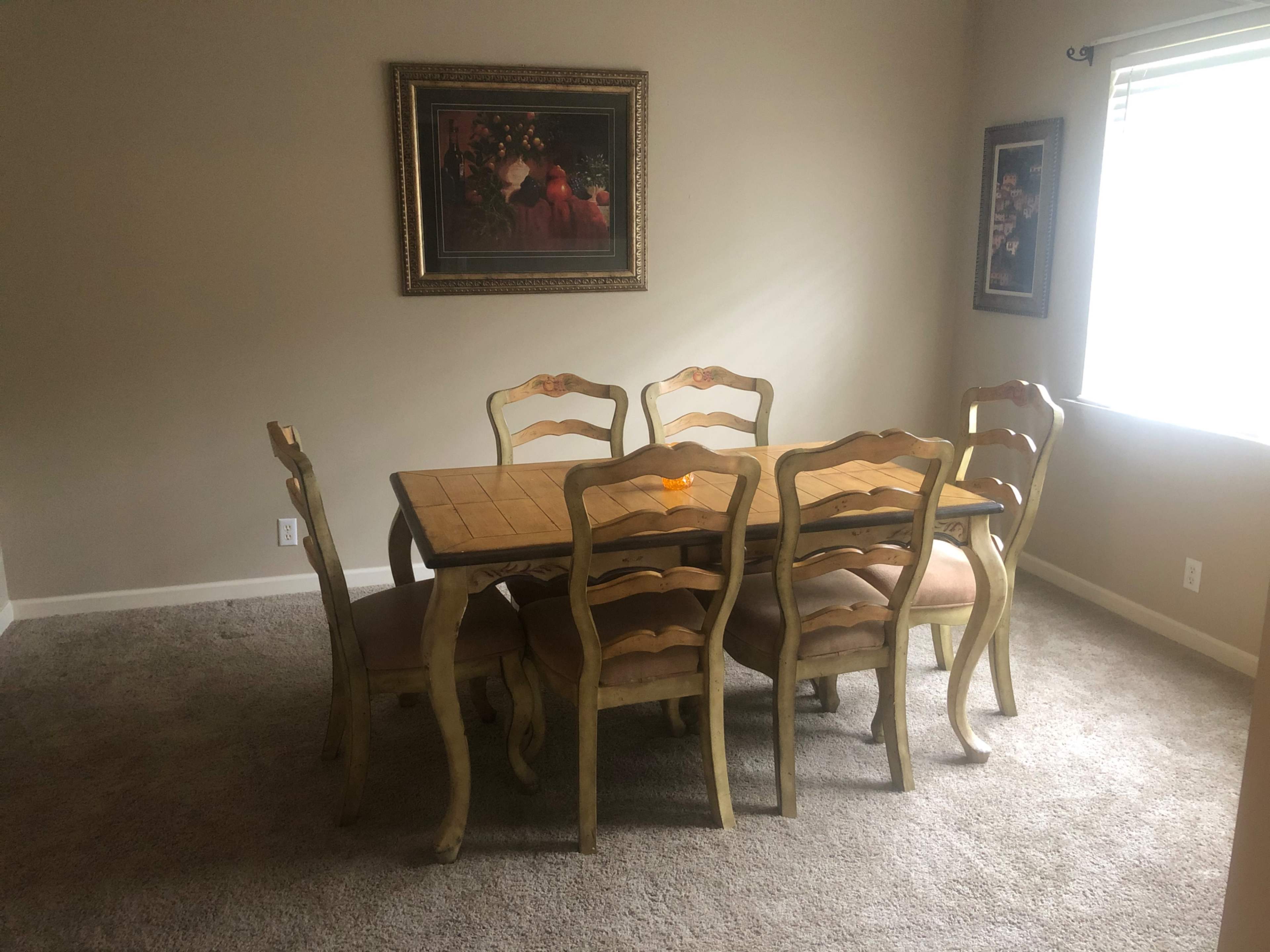 A wooden dining table surrounded by six chairs is positioned in a carpeted room with two framed pictures on the walls and a window that lets in natural light.