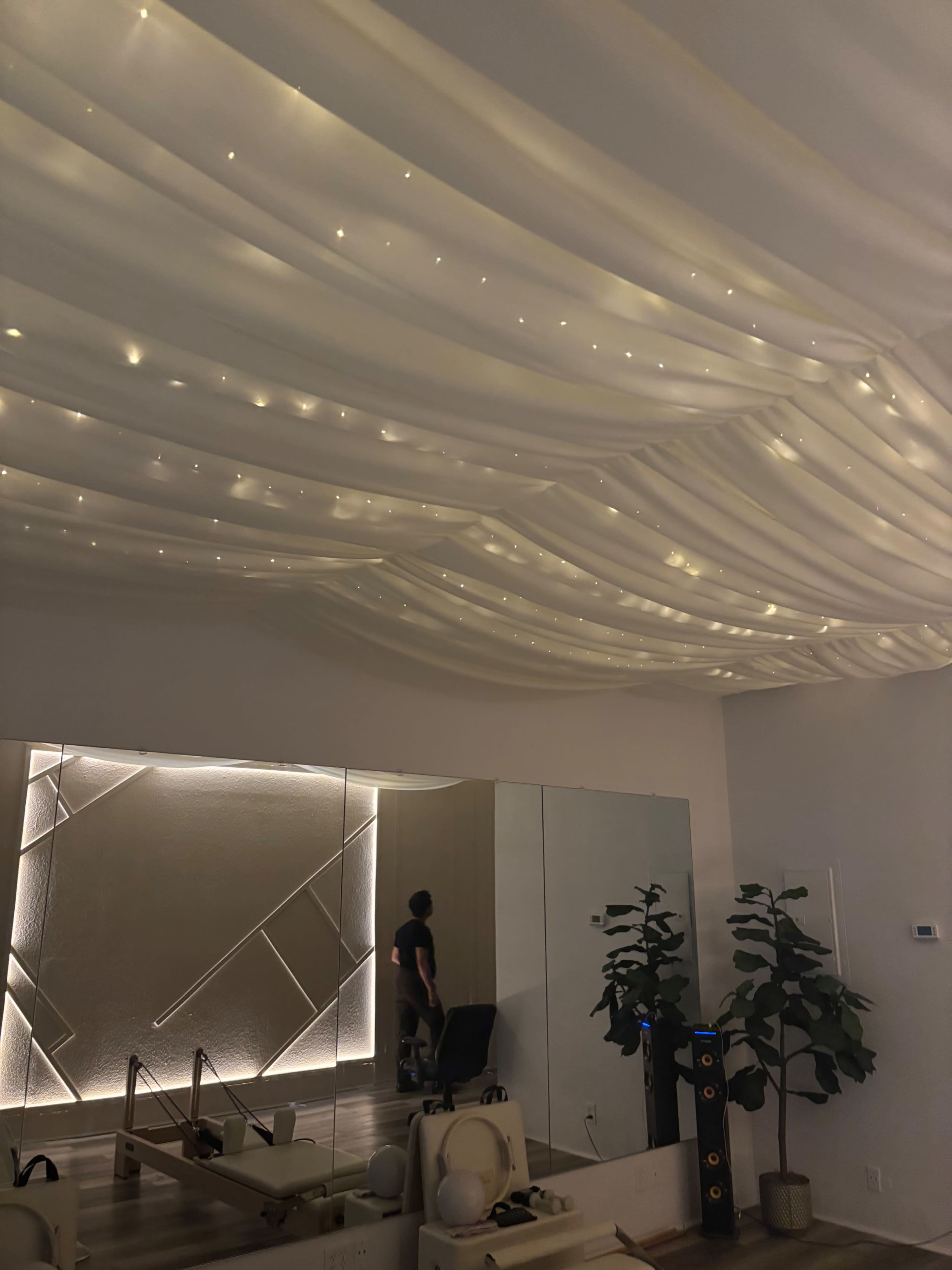The image shows a softly lit room with a draped ceiling, string lights, a large mirror, and a reformer pilates machine in the foreground.