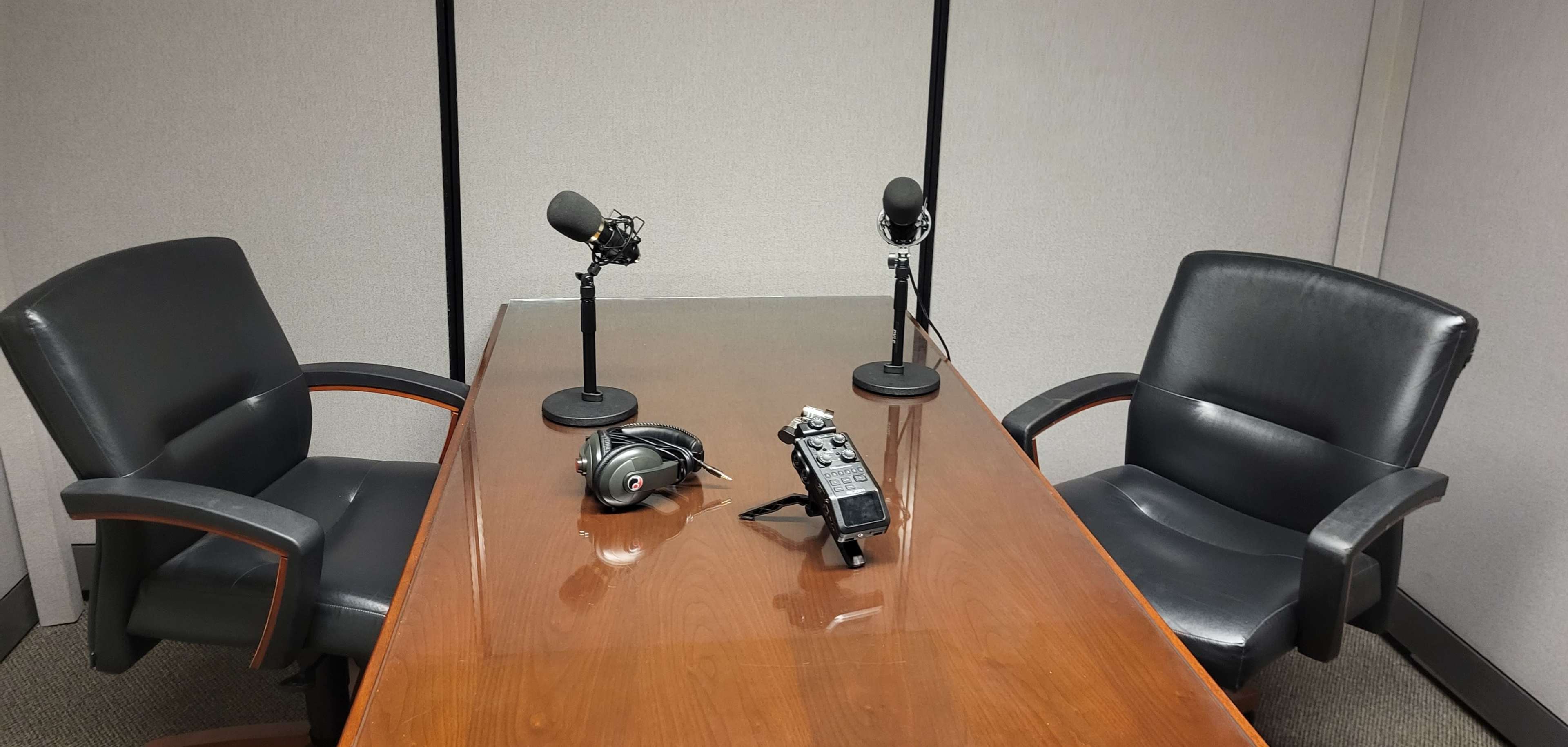 A conference room is set up with two microphones on stands, a recording device, and two black chairs around a wooden table.