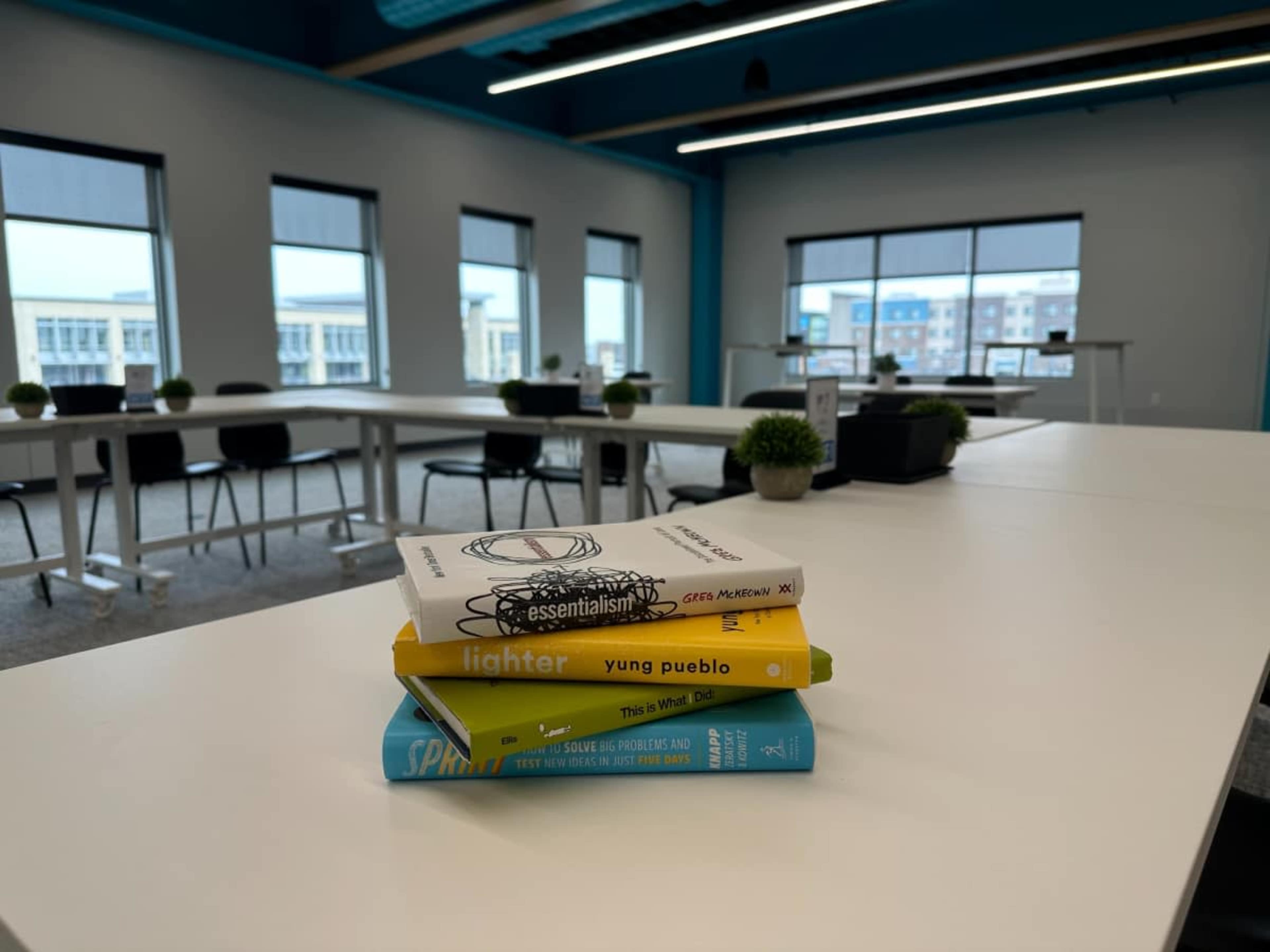 A stack of four books sits on a table in a modern office space with large windows and minimal furnishings.