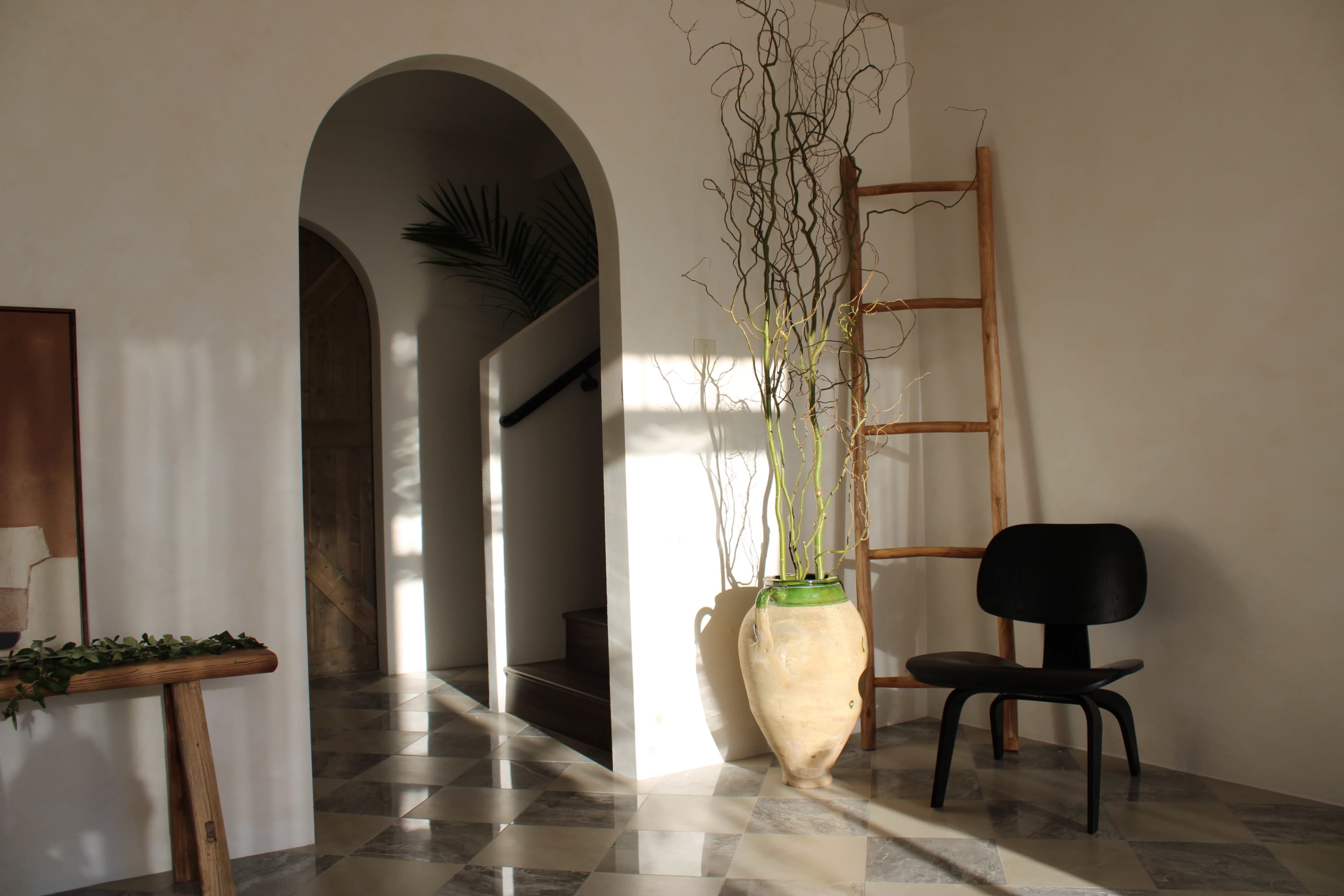 A minimalist interior with a wooden ladder, a black chair, a large vase with branches, and a staircase leading to a doorway.