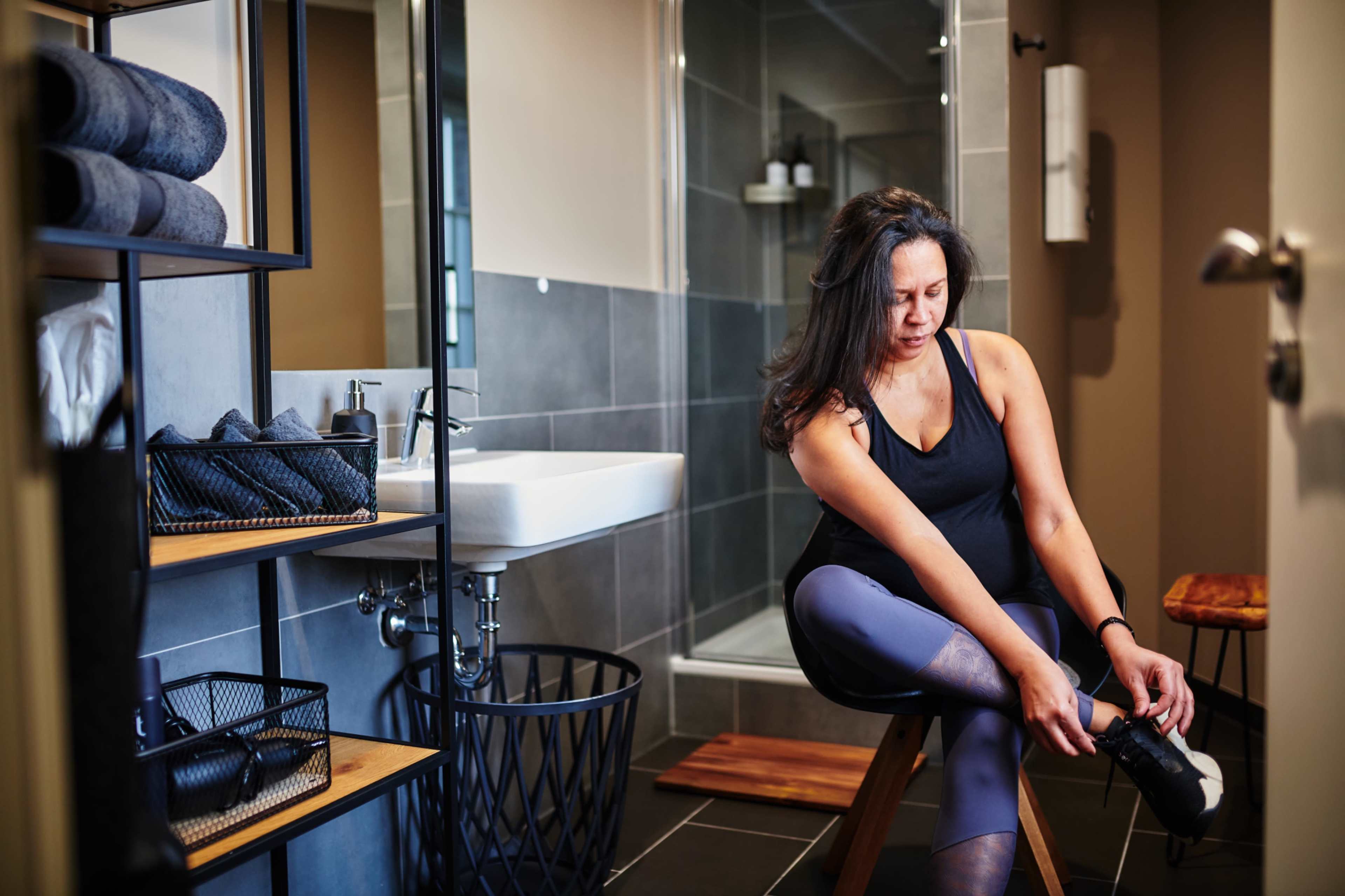 A woman is sitting on a wooden stool in a bathroom, putting on her shoes while surrounded by neatly arranged towels and toiletries.