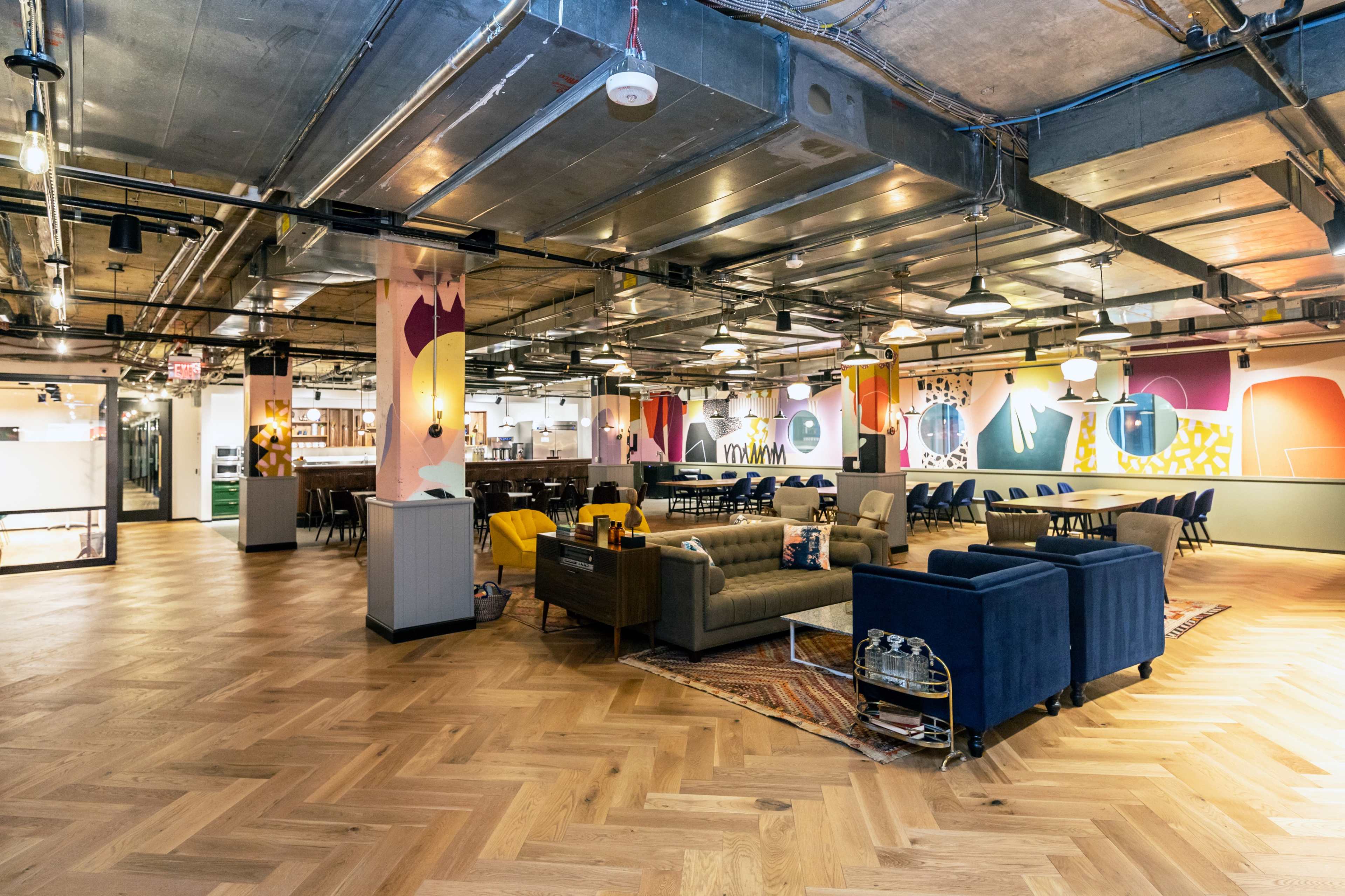 The image shows a spacious, modern interior of a communal workspace with wooden flooring, various seating areas, and colorful wall art.