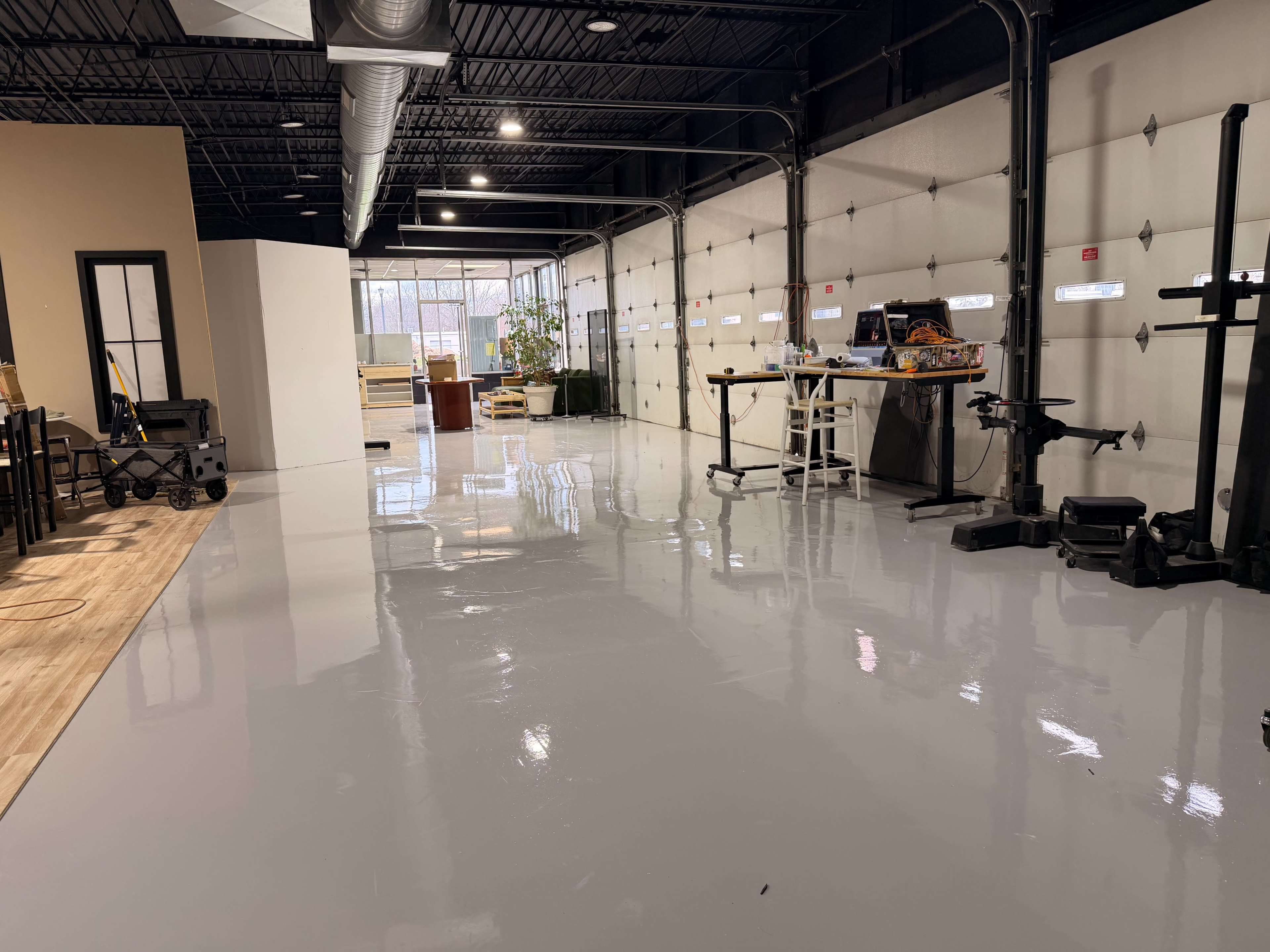 The image shows a large, polished concrete floor inside a well-lit industrial space, with garage doors on one side and various furniture and equipment scattered throughout.