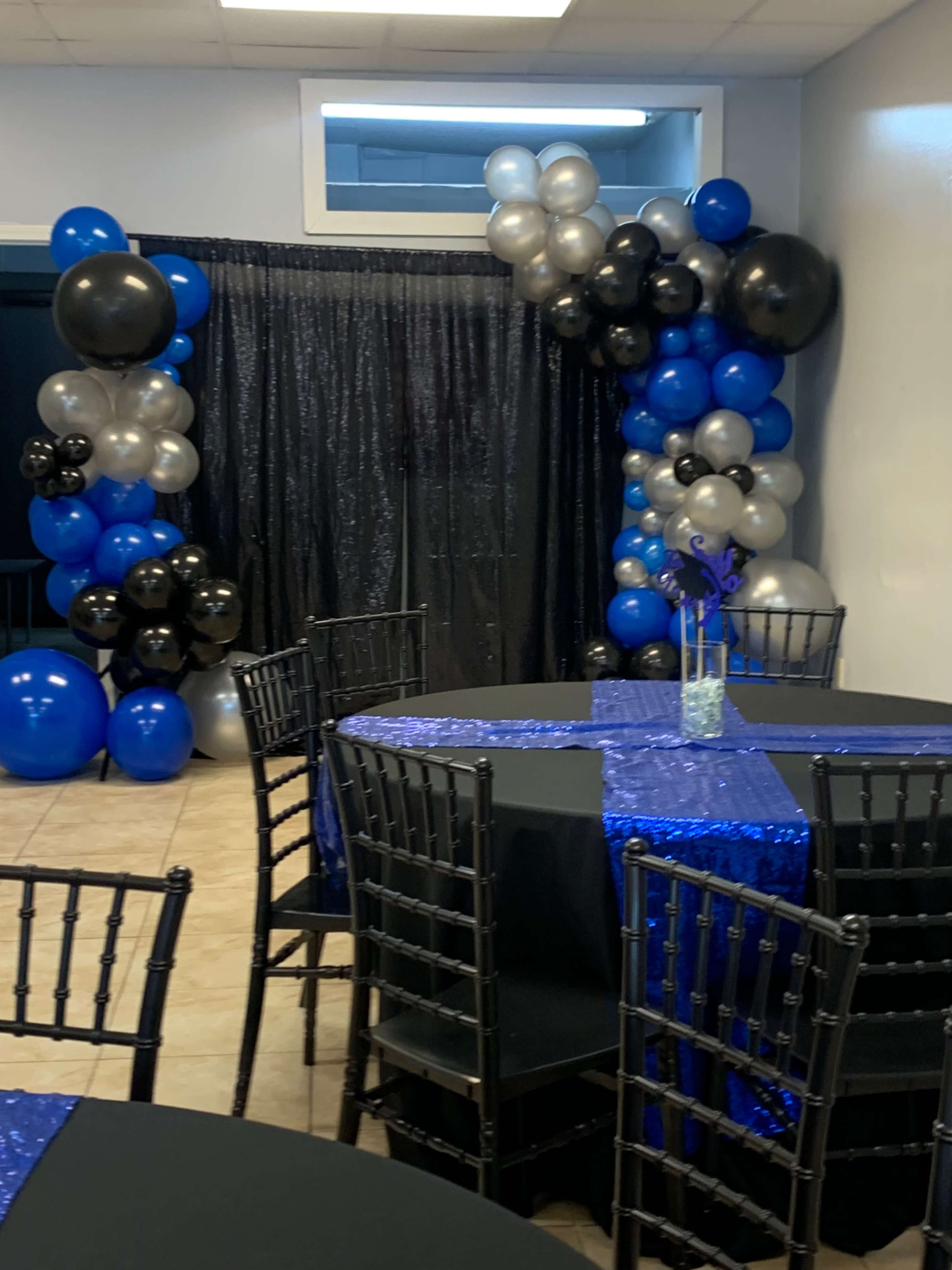 A decorated event space featuring black tables with blue tablecloths, surrounded by black, blue, and silver balloon arrangements and a black curtain backdrop.
