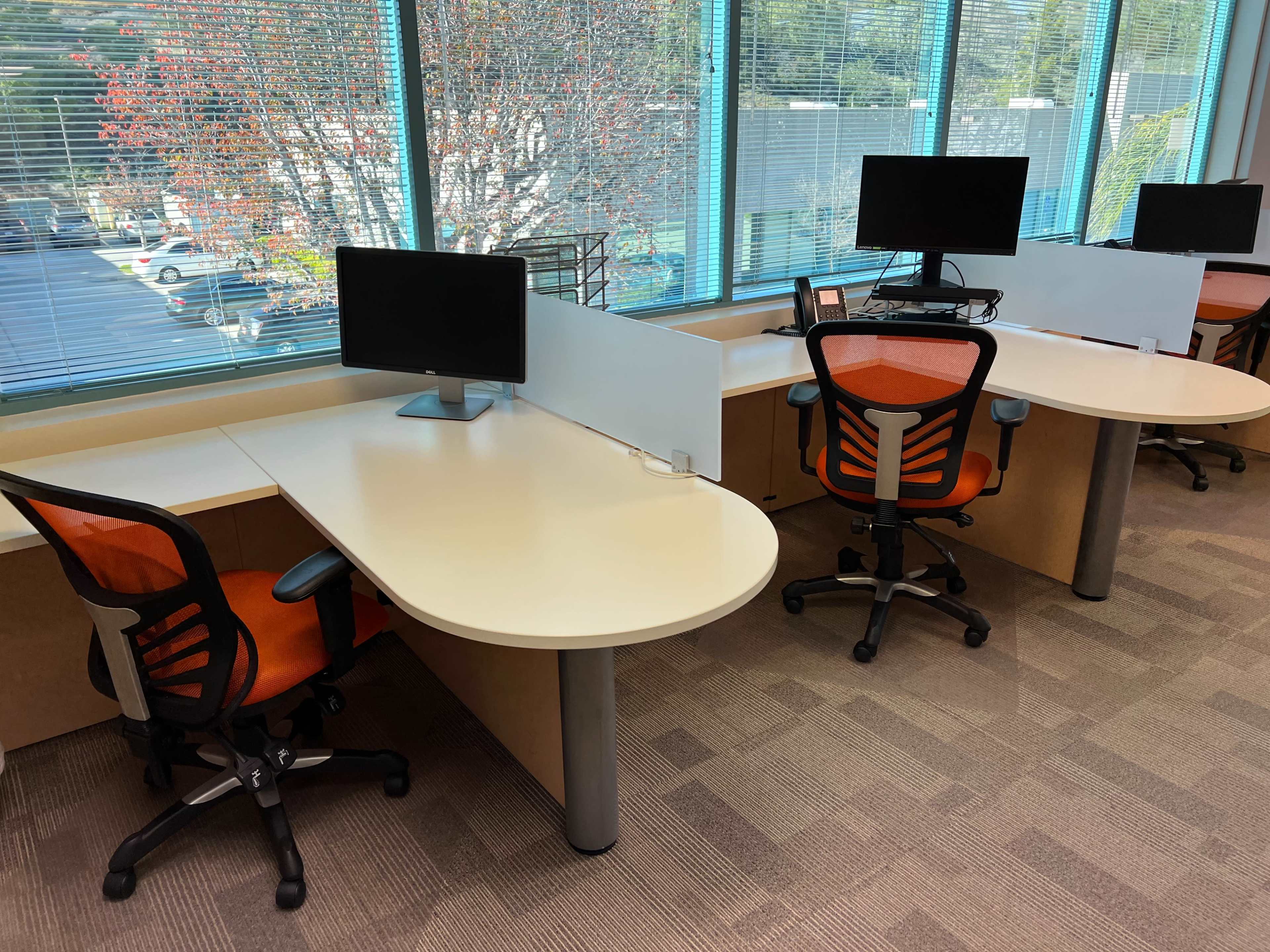 A brightly lit office space with two workstations, each equipped with a computer monitor and an orange office chair, separated by a translucent divider.