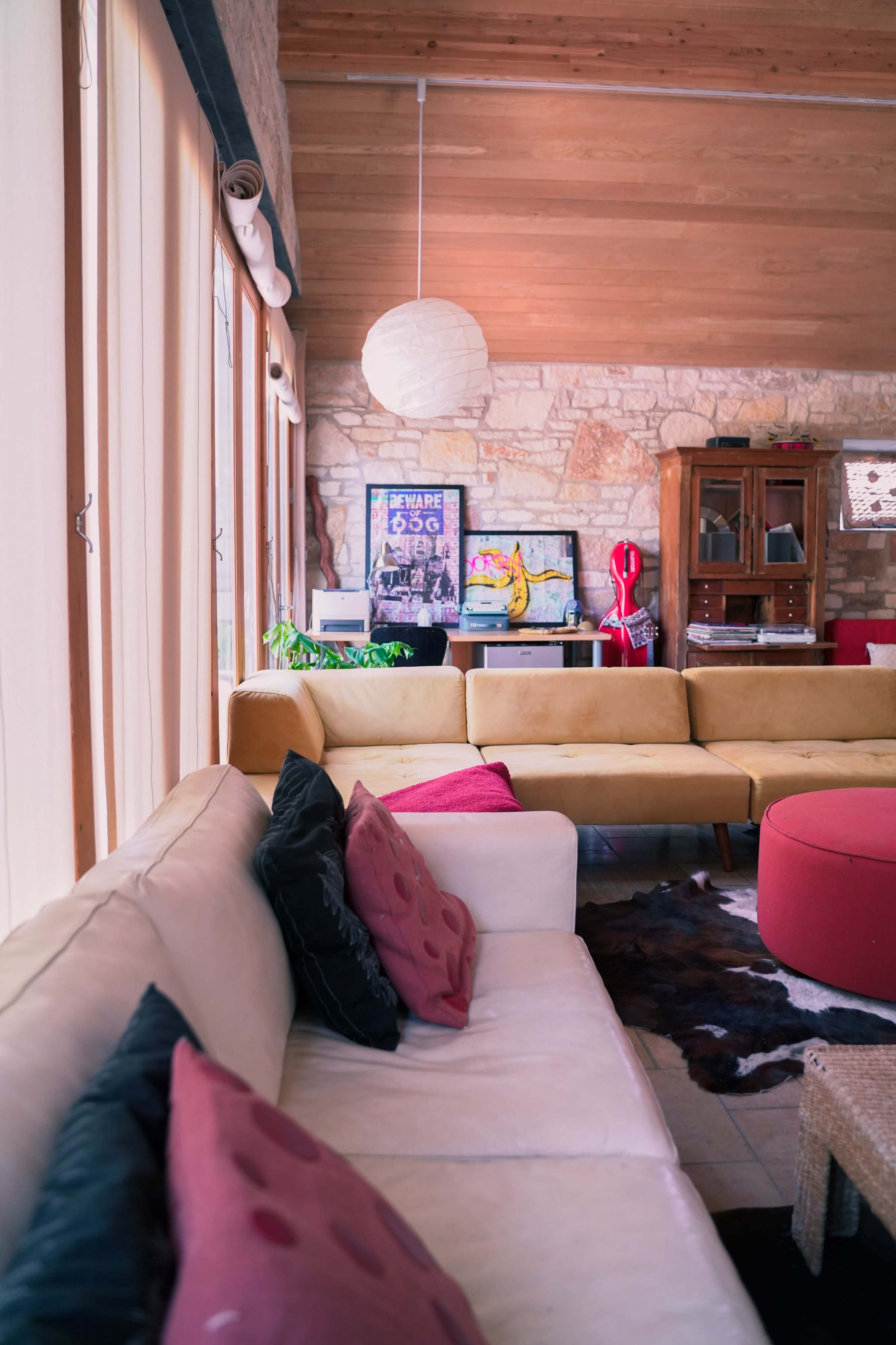 A cozy living room with a beige sofa, colorful cushions, a round red ottoman, and a mix of modern and rustic decor elements.