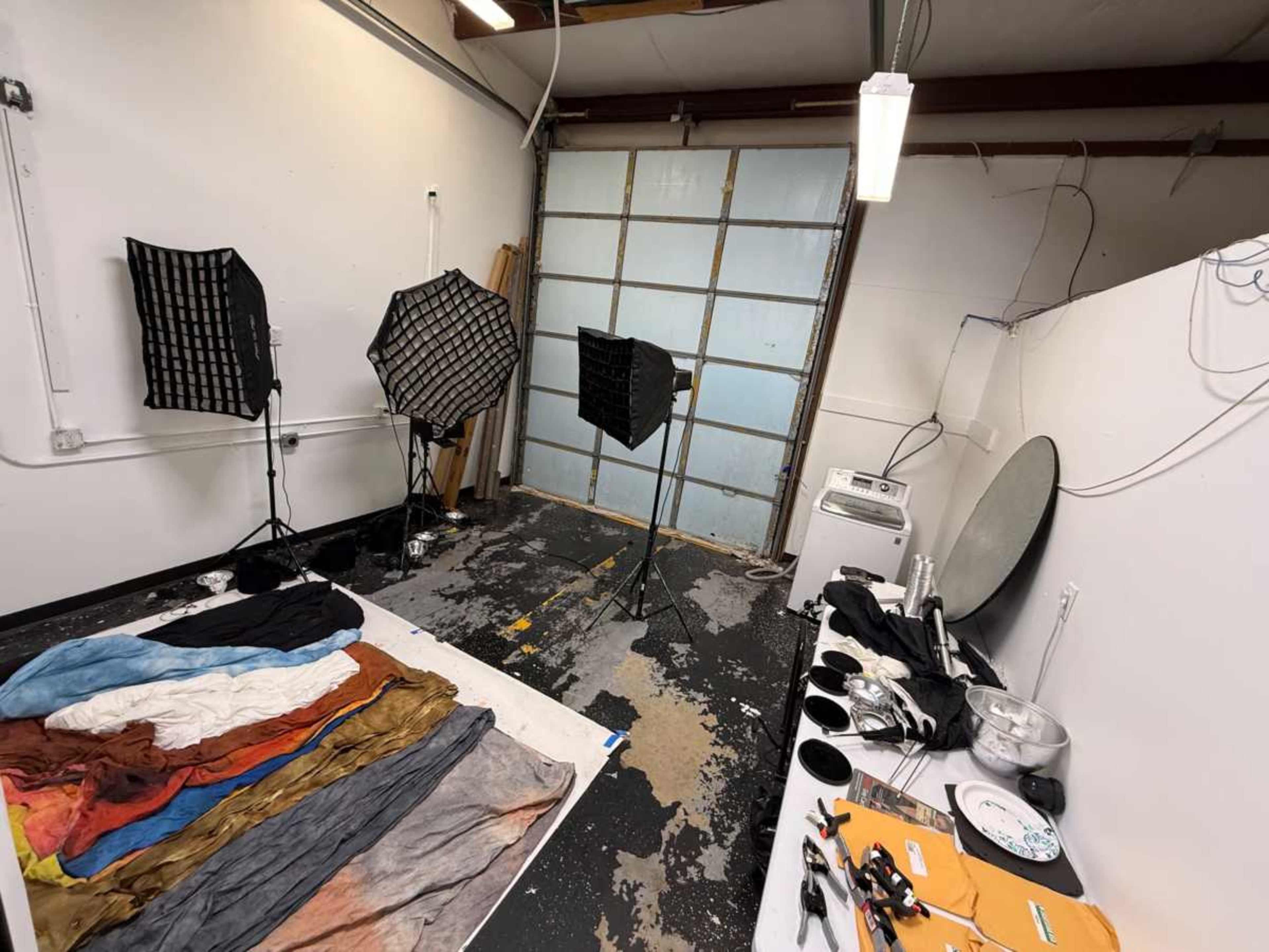 The image shows a photography studio with lighting equipment, backdrops, and various props scattered across the floor.