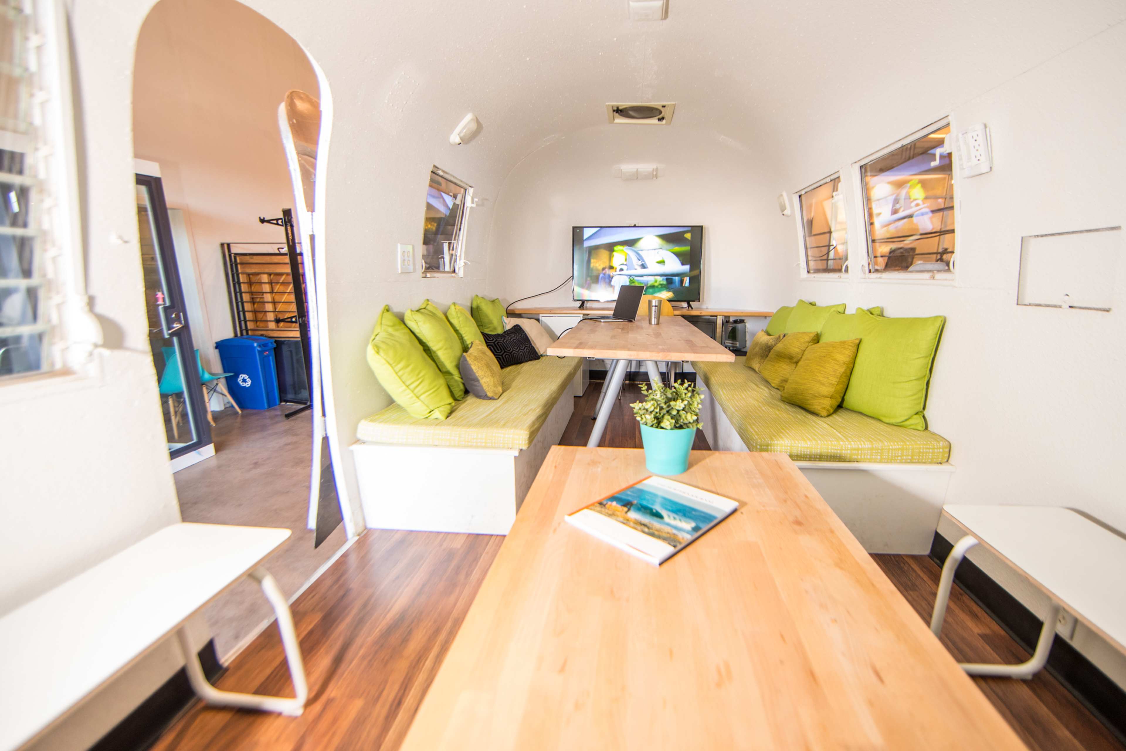 The interior of a modern trailer features a long wooden table surrounded by cushioned seating and a television mounted on the wall.