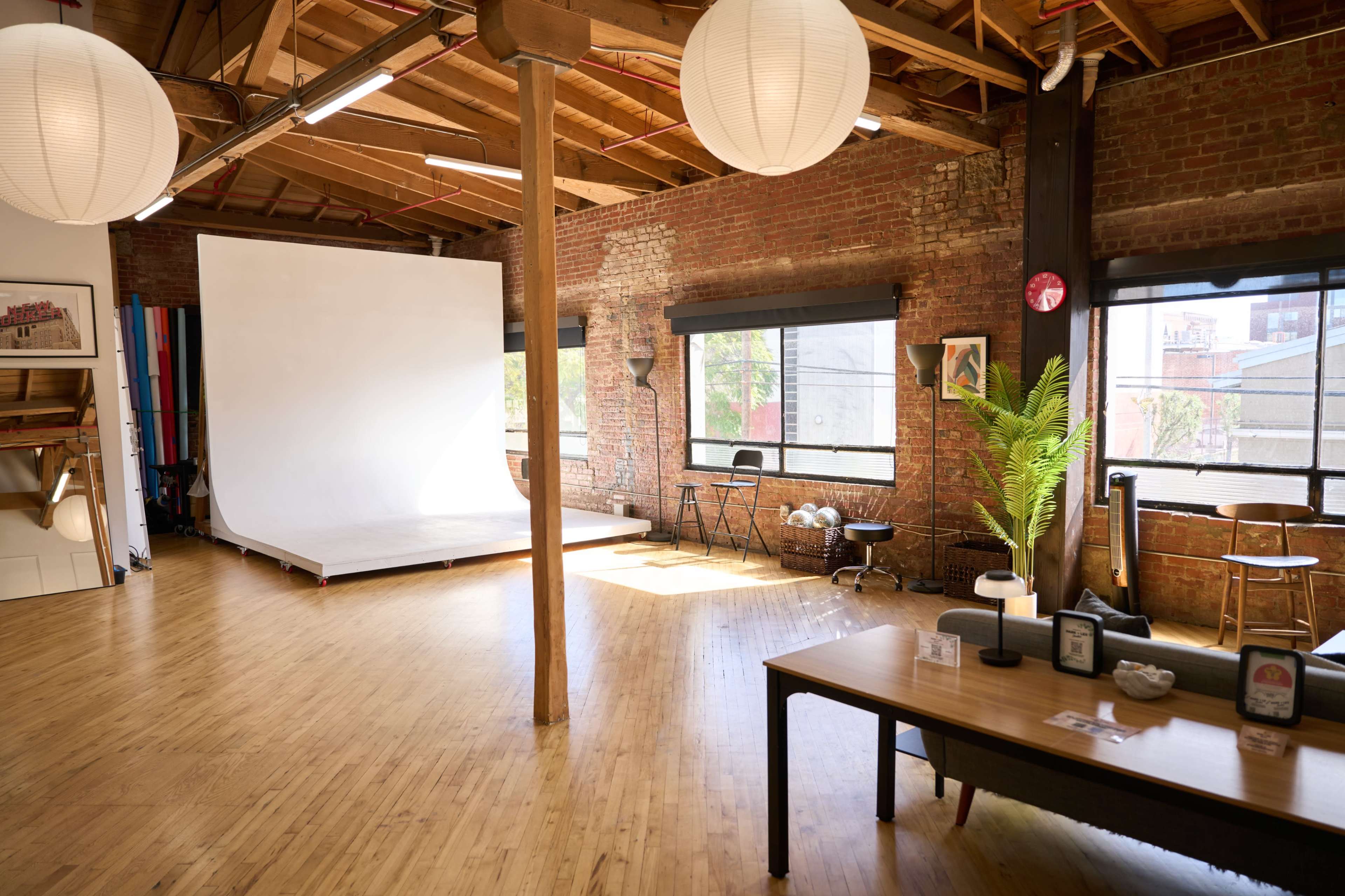 The image shows a spacious, well-lit studio with wooden flooring, large windows, a white backdrop on a raised platform, and a table with decorative items.