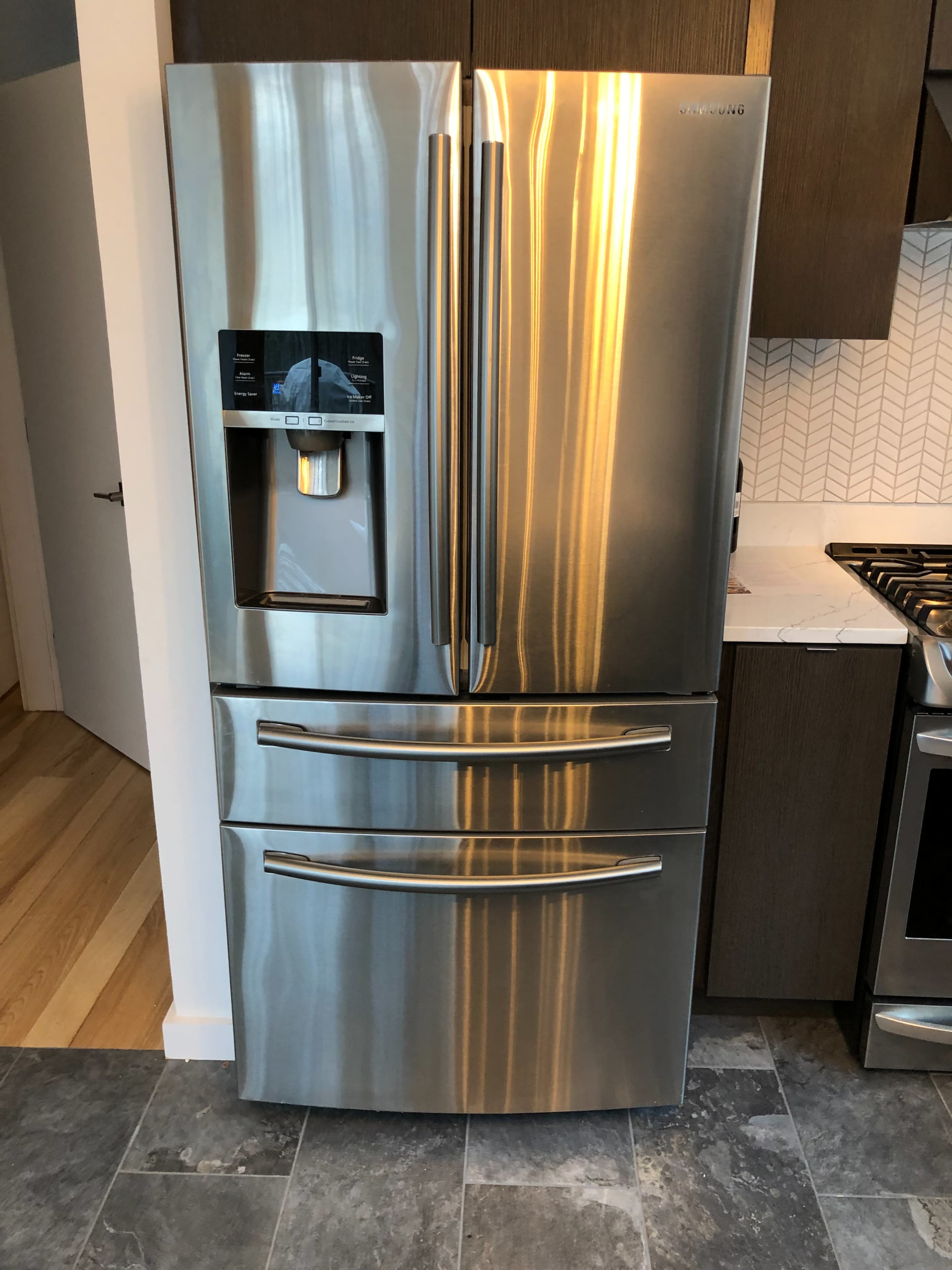 A stainless steel refrigerator with a water and ice dispenser is positioned in a modern kitchen with dark cabinets and a tiled backsplash.