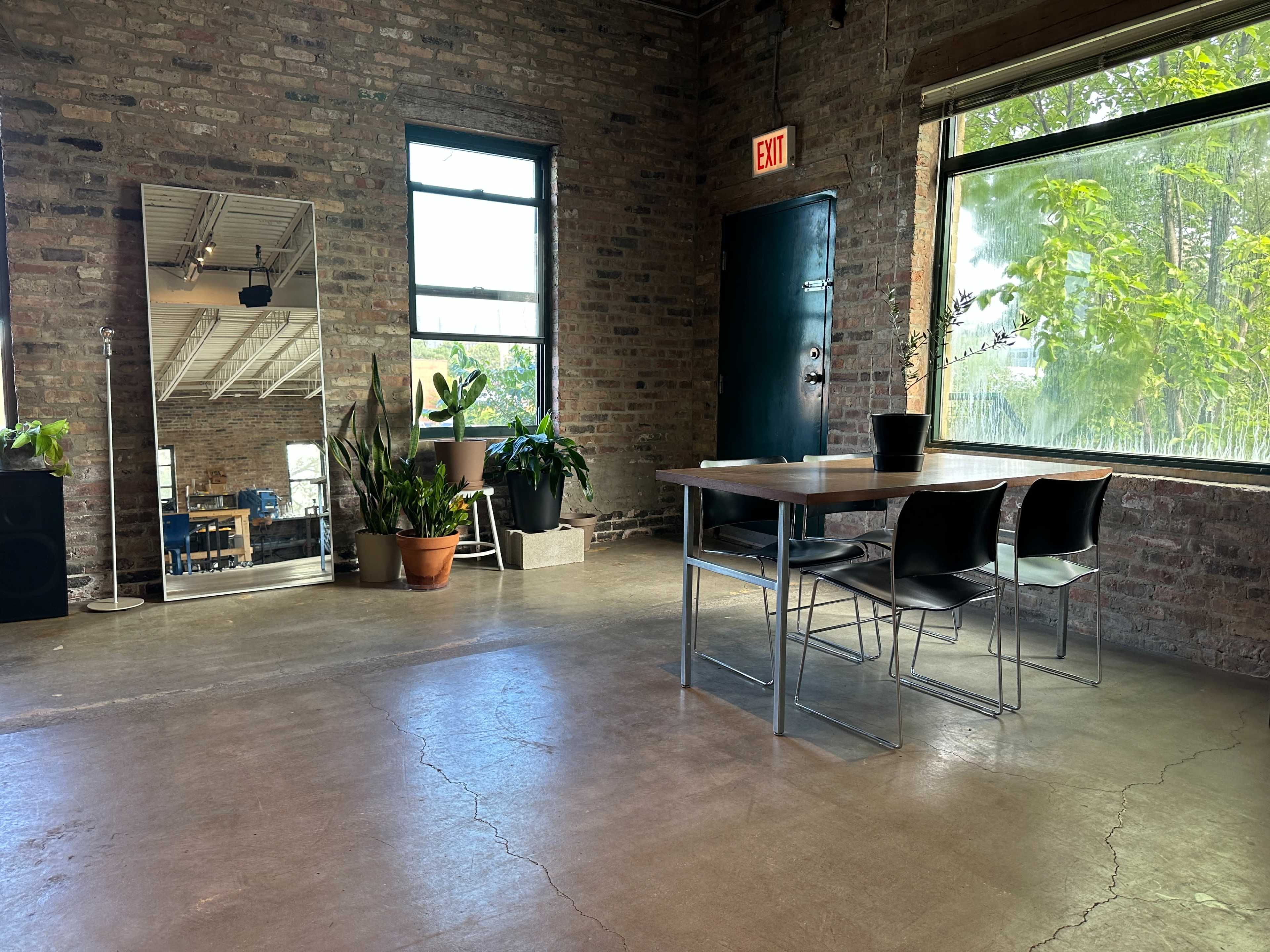 The scene shows a spacious room with exposed brick walls, a table surrounded by four black chairs, and large windows allowing natural light to enter, along with several potted plants.