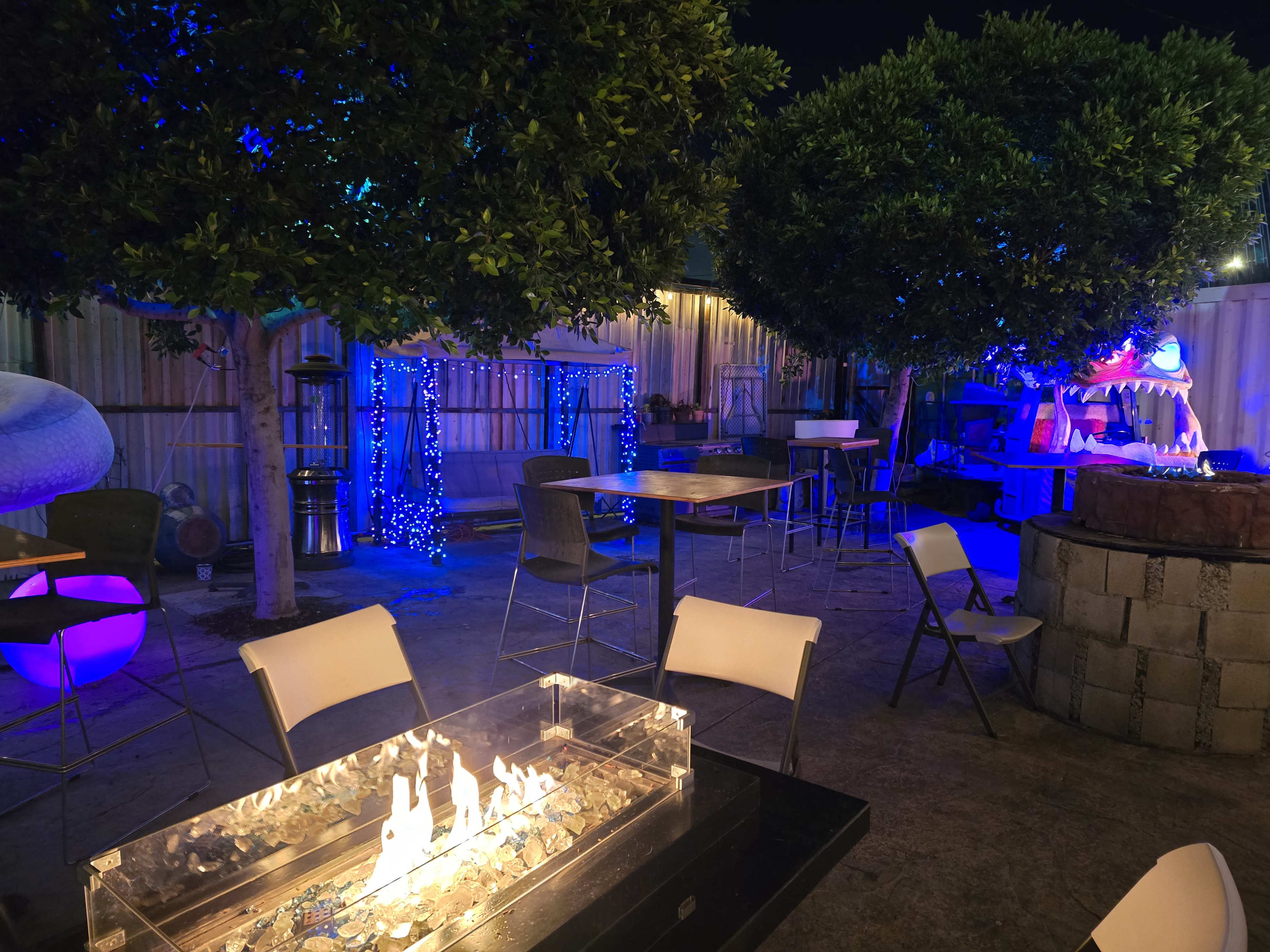 The image shows an outdoor seating area at night, featuring a fire pit, tables, and decorative trees illuminated by blue lights.