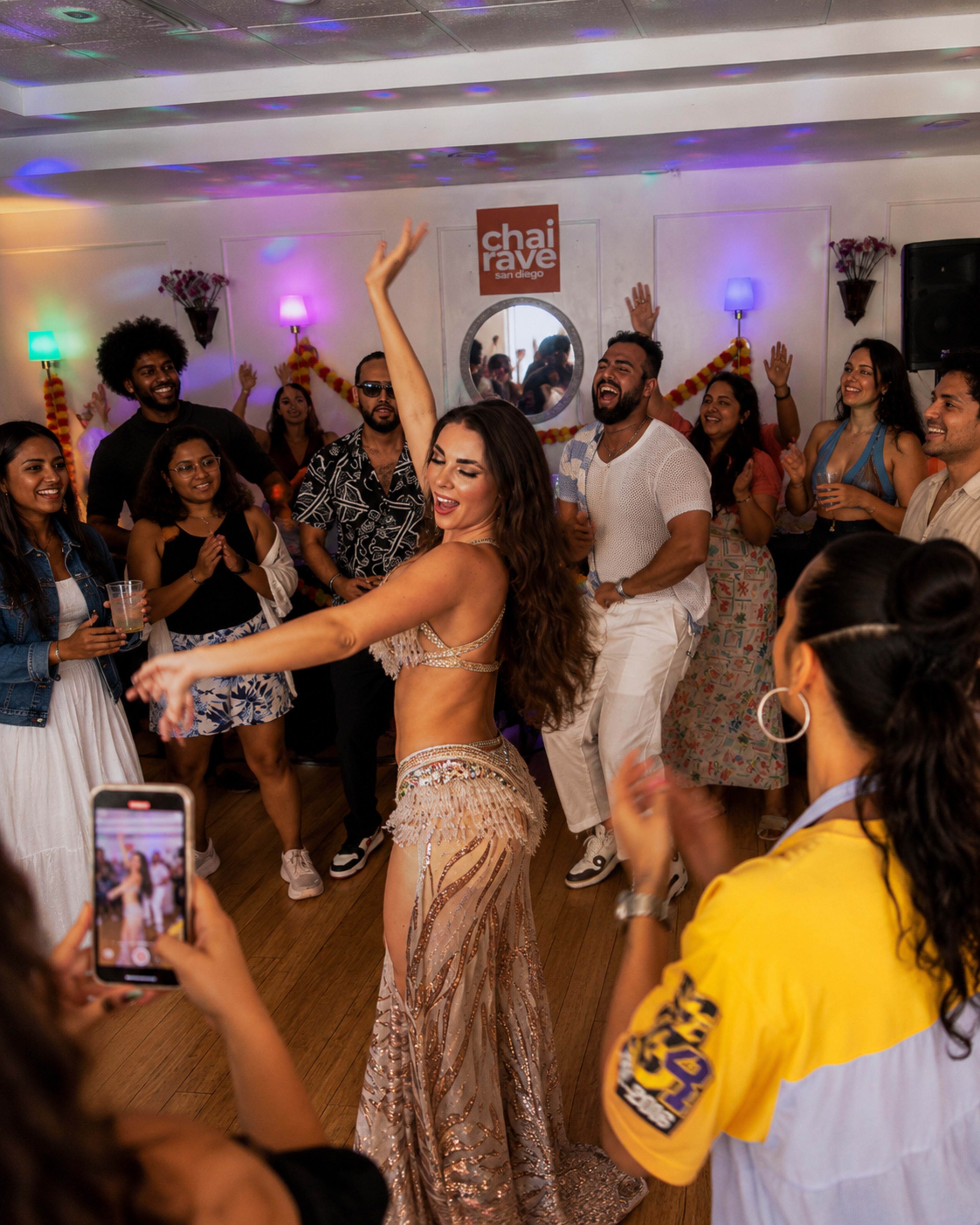 A woman in a sparkling costume performs a dance in a lively room filled with people celebrating.