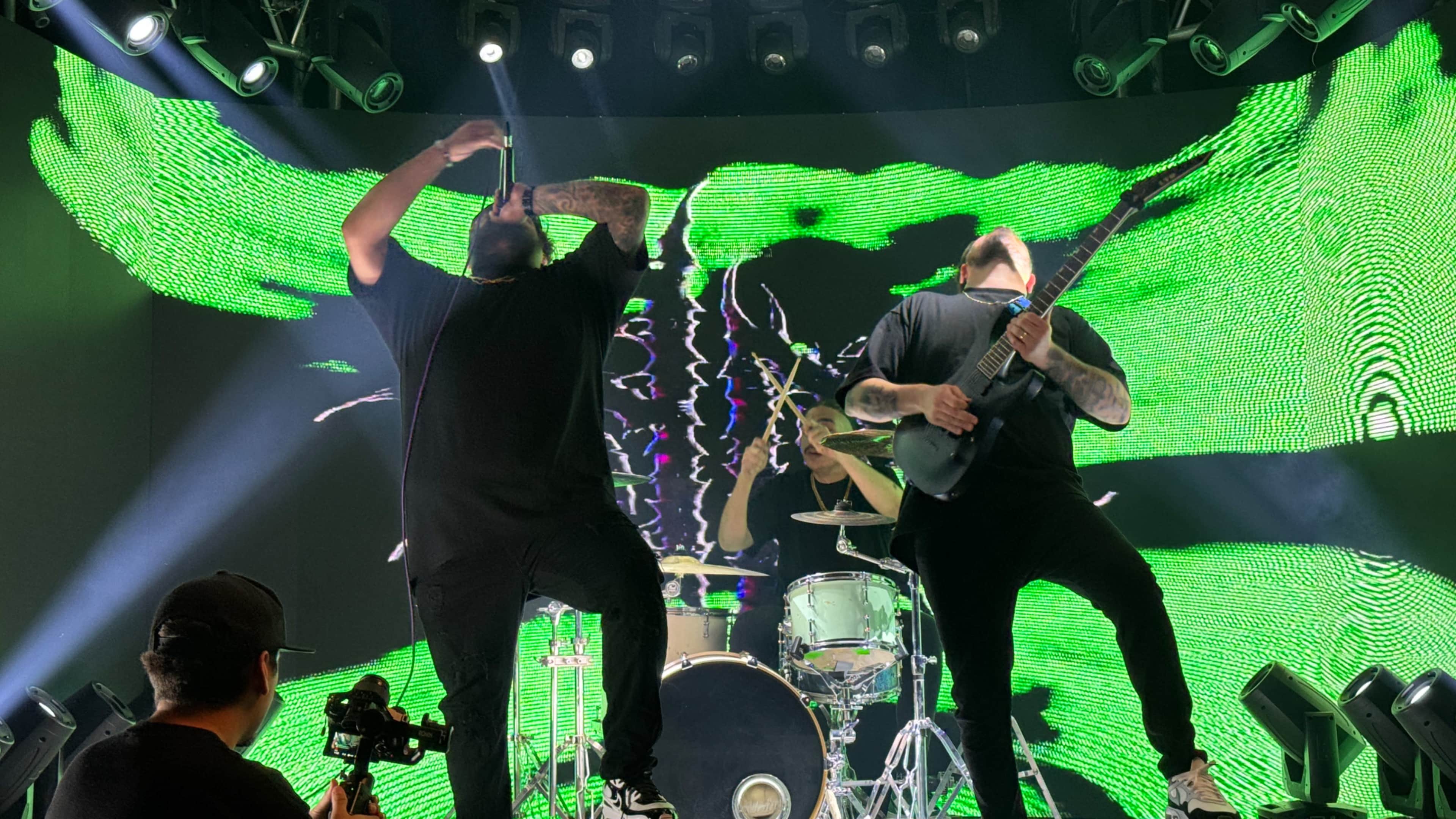 A band performs on stage with vibrant green visuals in the background, featuring a vocalist, guitarist, and drummer.