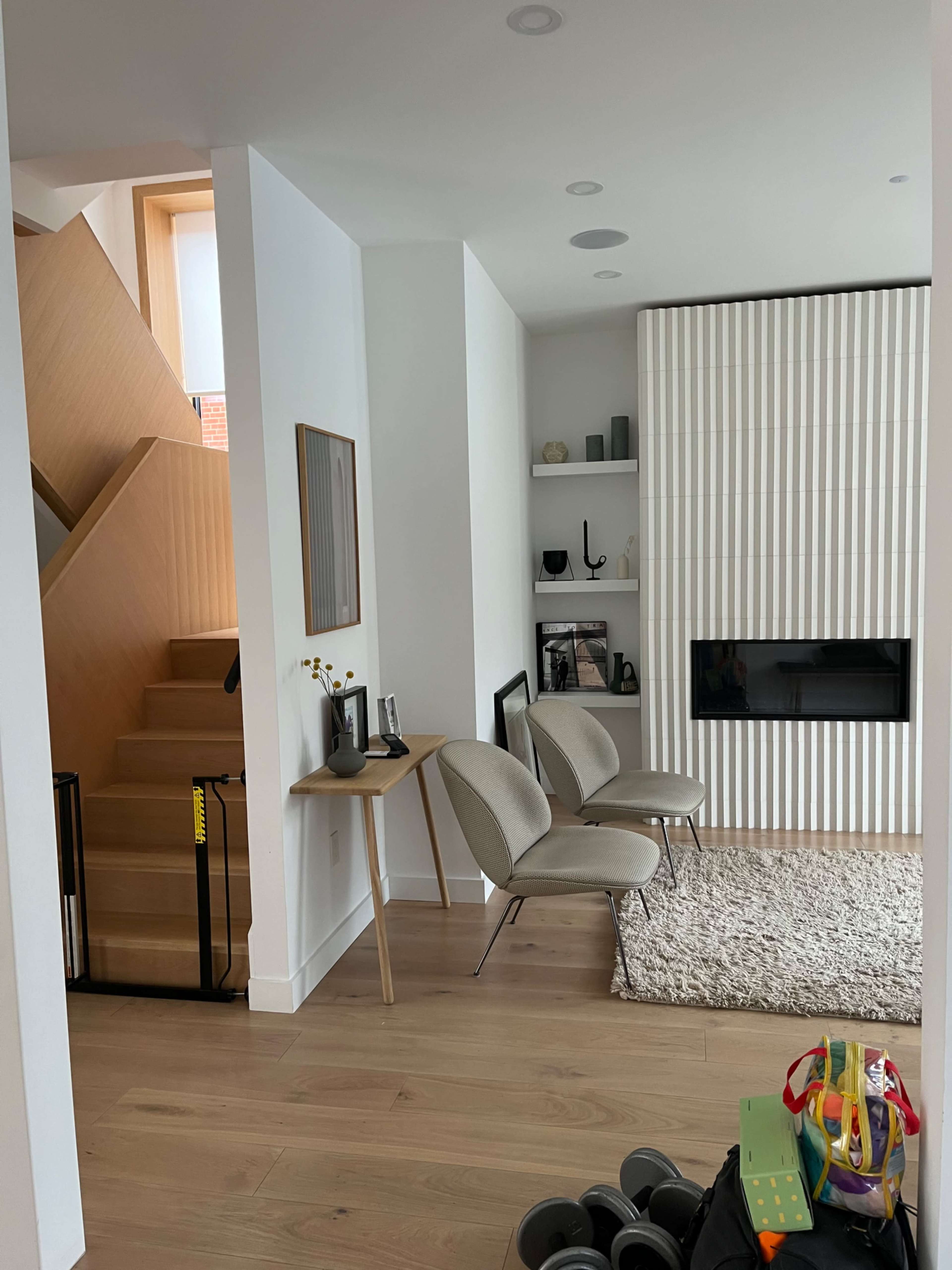 A modern living area with two chairs, a table, a staircase in the background, and a wall-mounted fireplace.