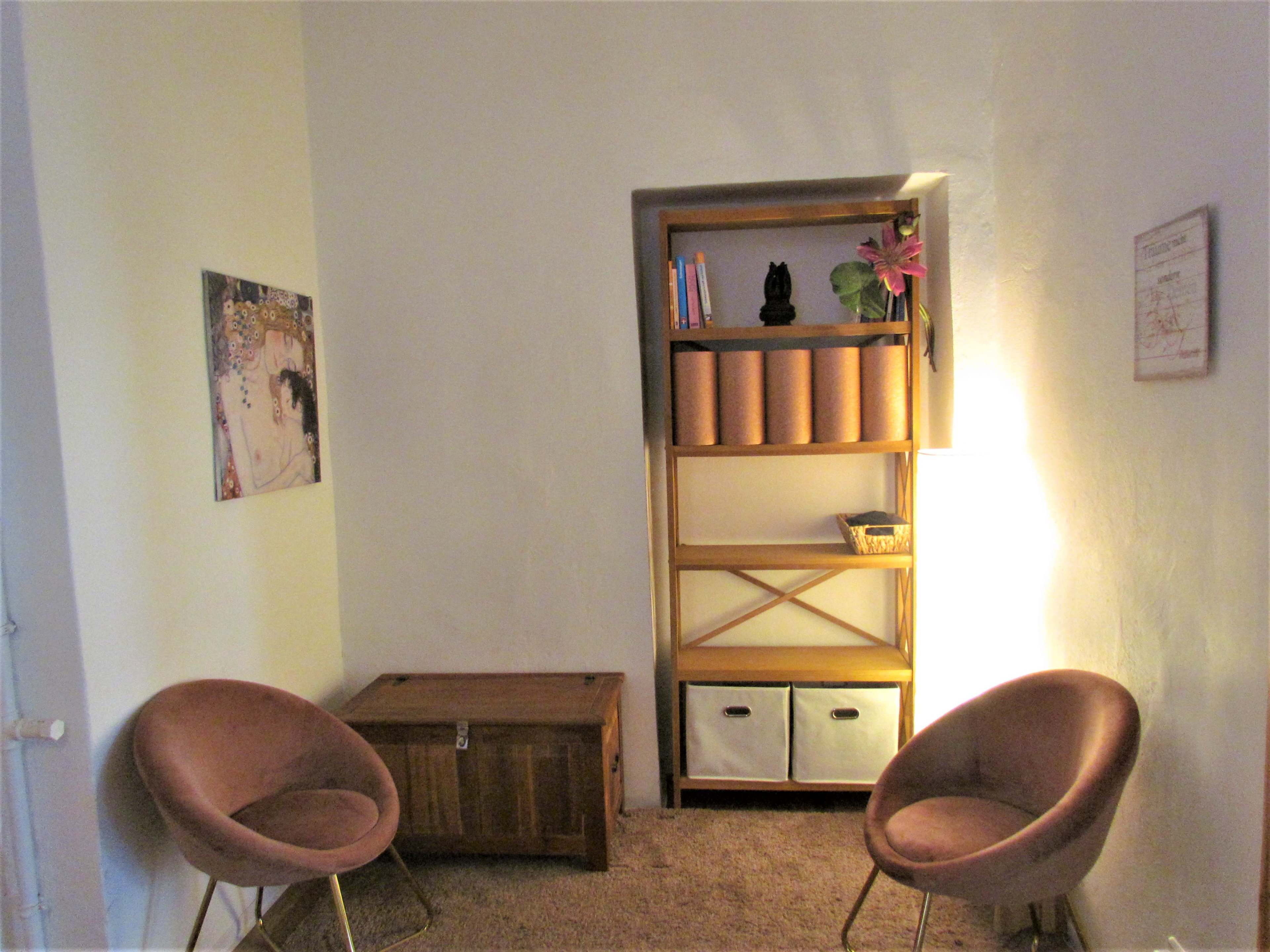 A small room features two chairs facing a wooden storage box and a shelf with books and decorative items, illuminated by a standing lamp.