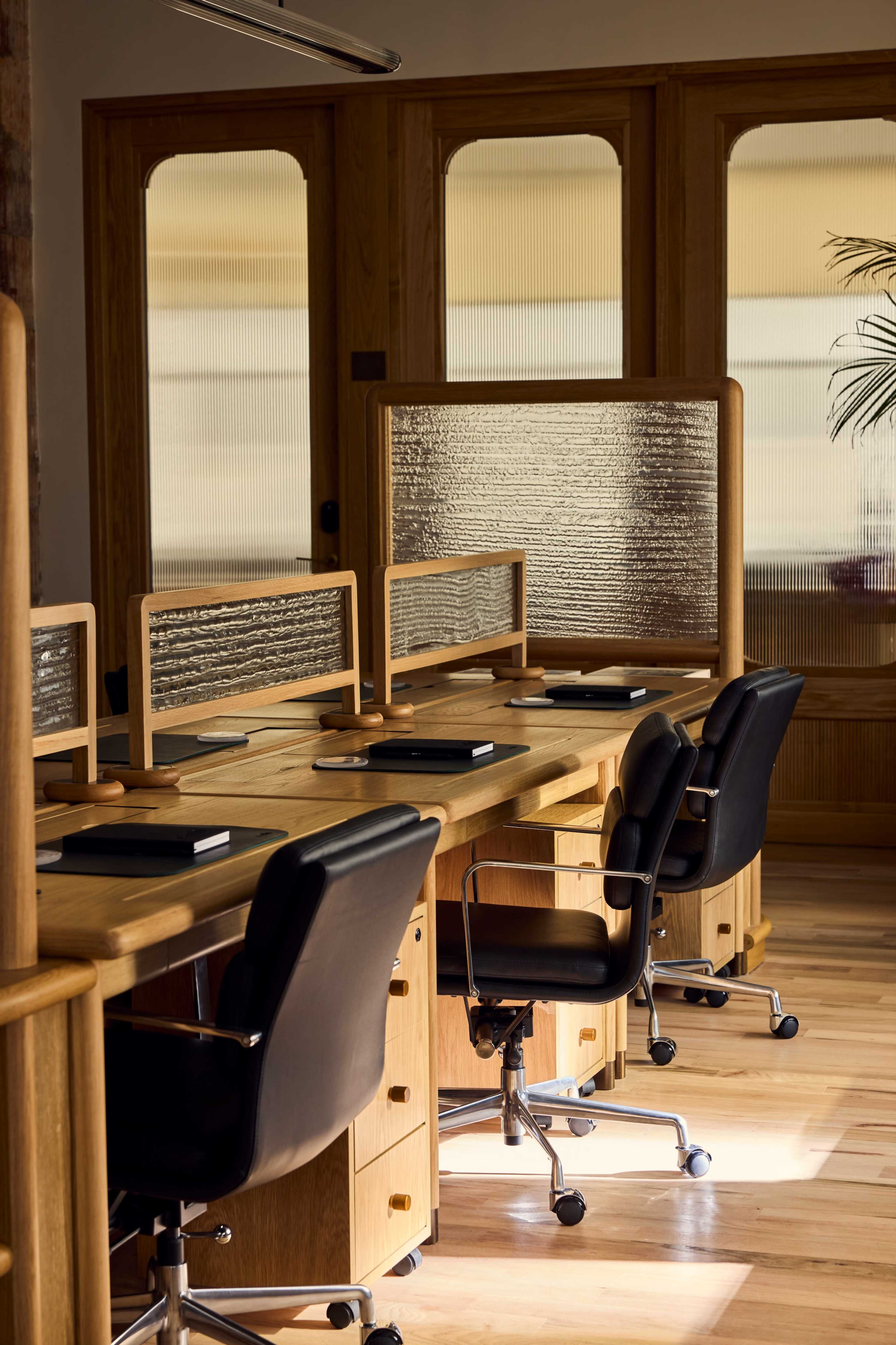 The image shows a modern office interior with wooden desks and black office chairs arranged for multiple workstations.