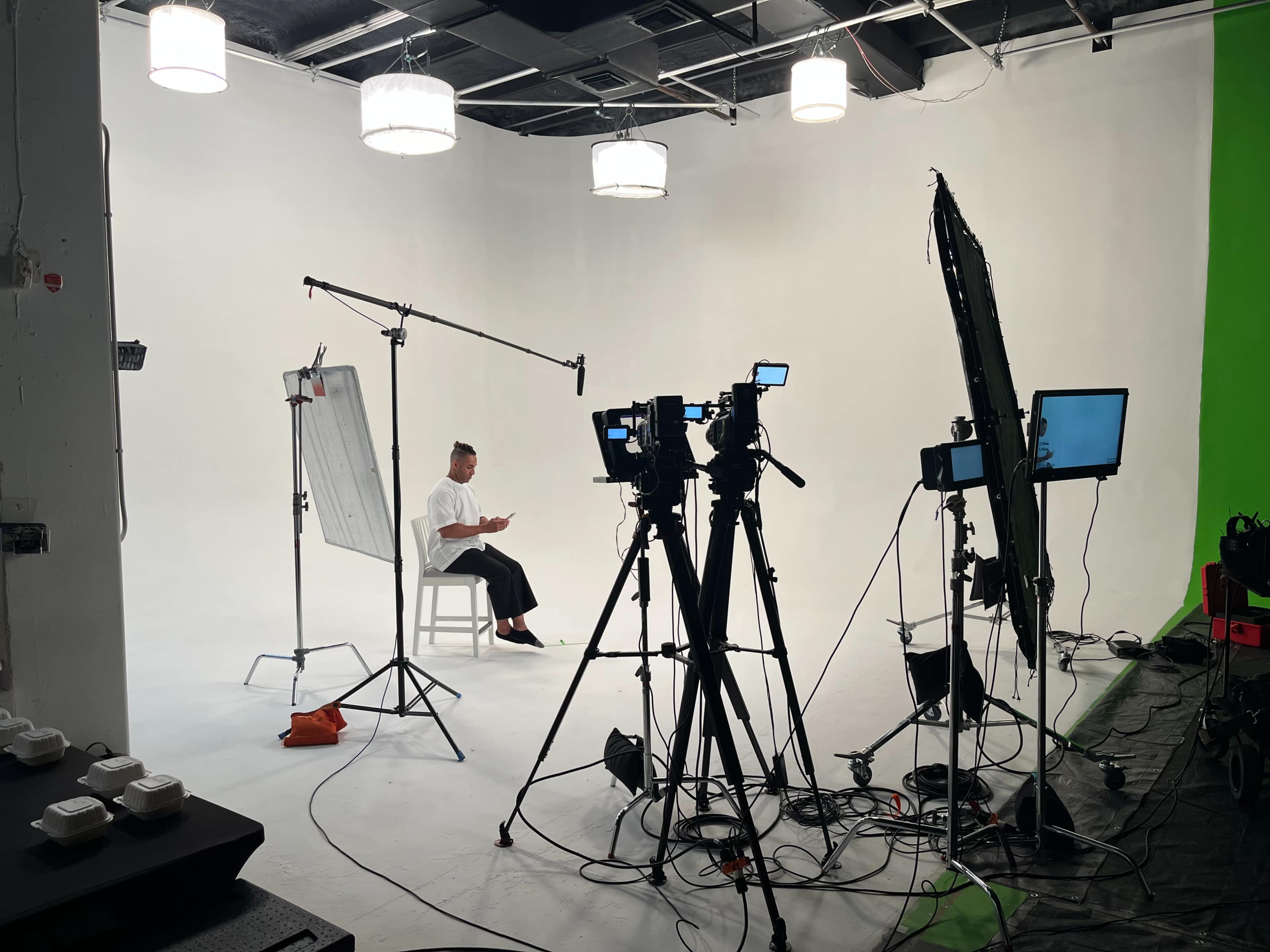A person is seated on a chair in a studio, surrounded by multiple cameras and equipment, with a white backdrop and lights overhead.