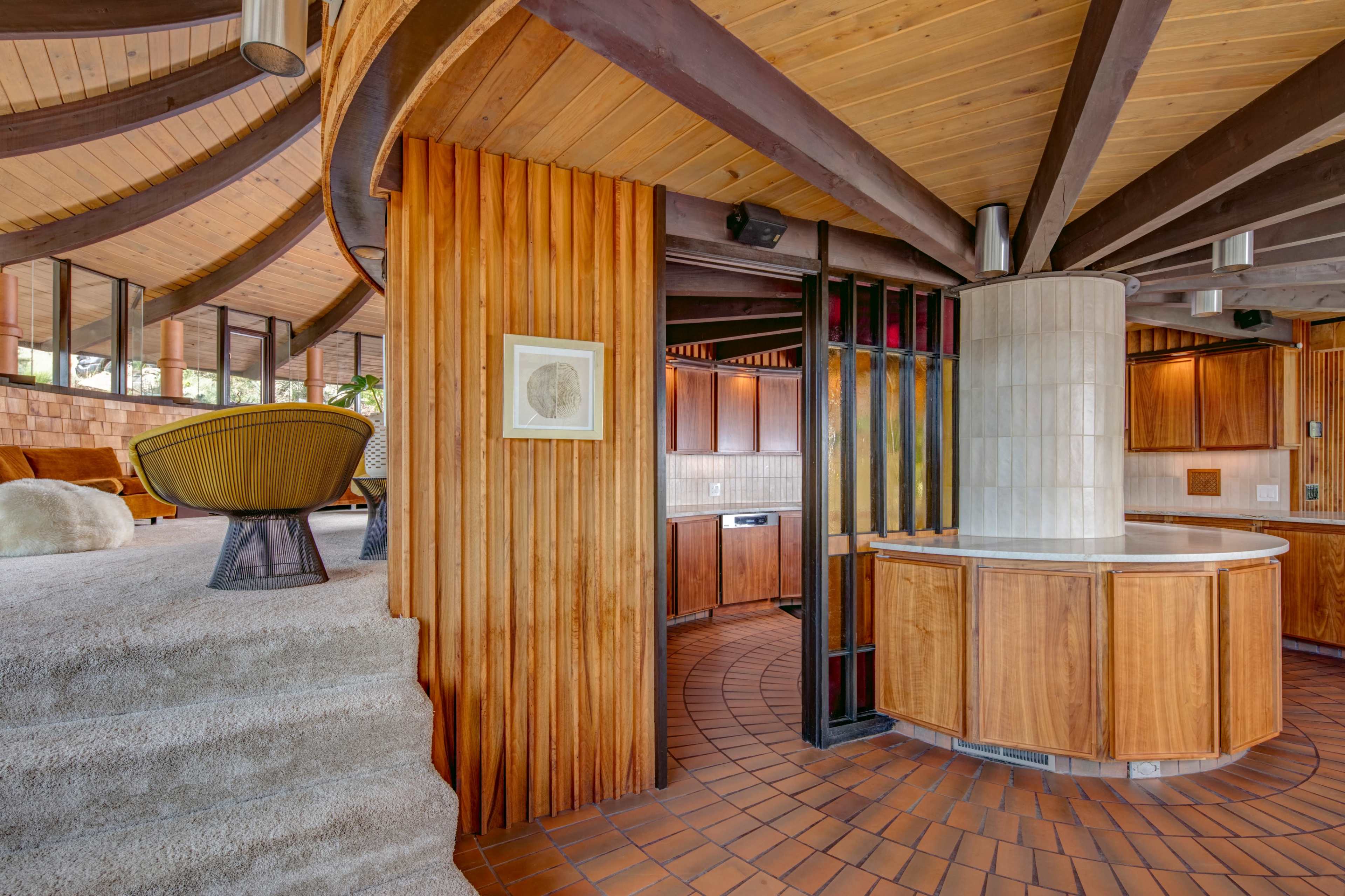 The image displays the interior of a modern home with curved wooden walls, an open layout, and distinctive architectural features, including a circular kitchen island and a staircase.