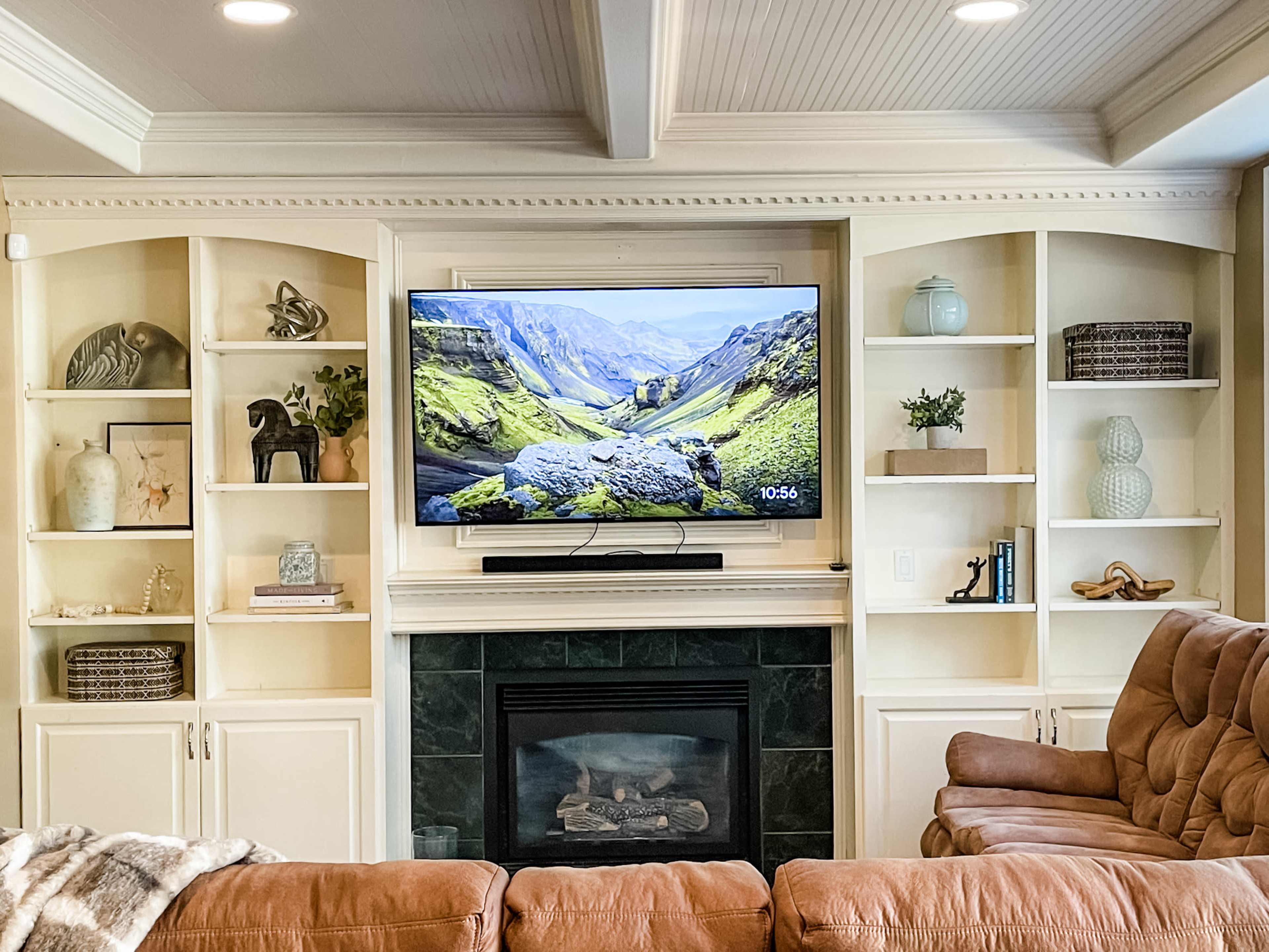 A flat-screen television is mounted above a fireplace in a room with built-in shelves featuring various decorative items and a brown sectional couch.