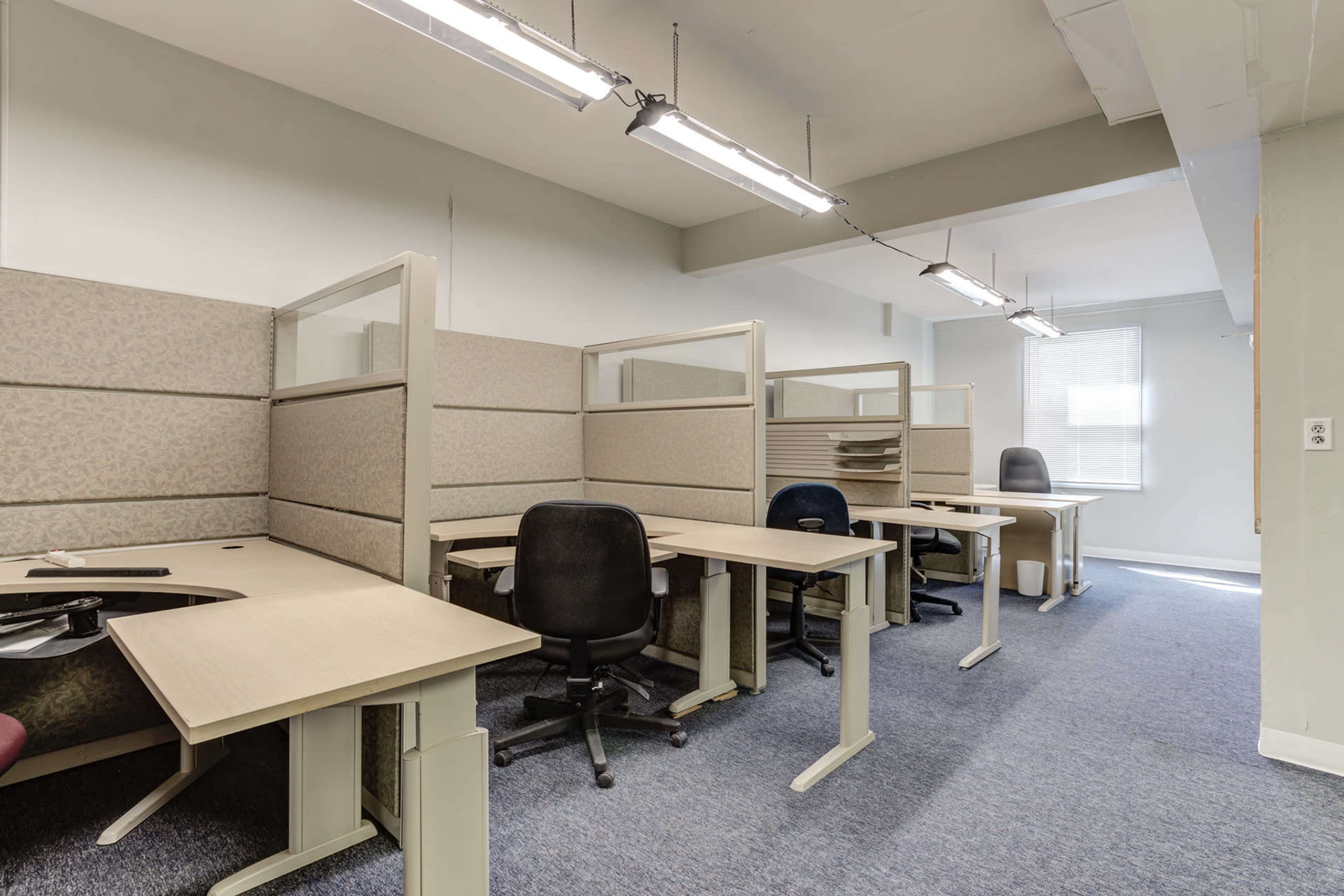 A row of empty office cubicles with desks and task chairs in a well-lit room.