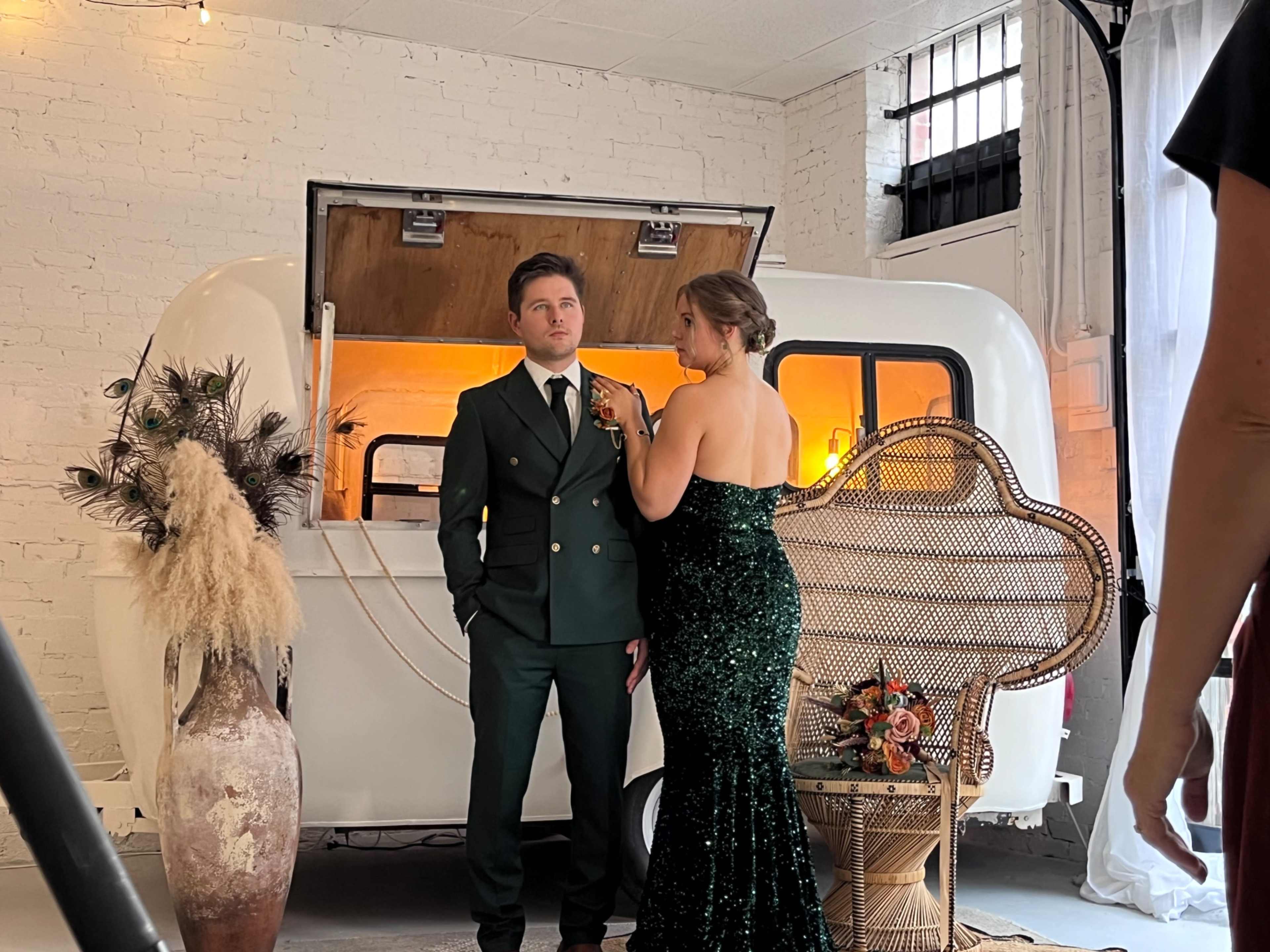 A man in a dark green suit stands beside a woman in a sparkling green dress, set against a vintage white camper trailer in a well-lit room.