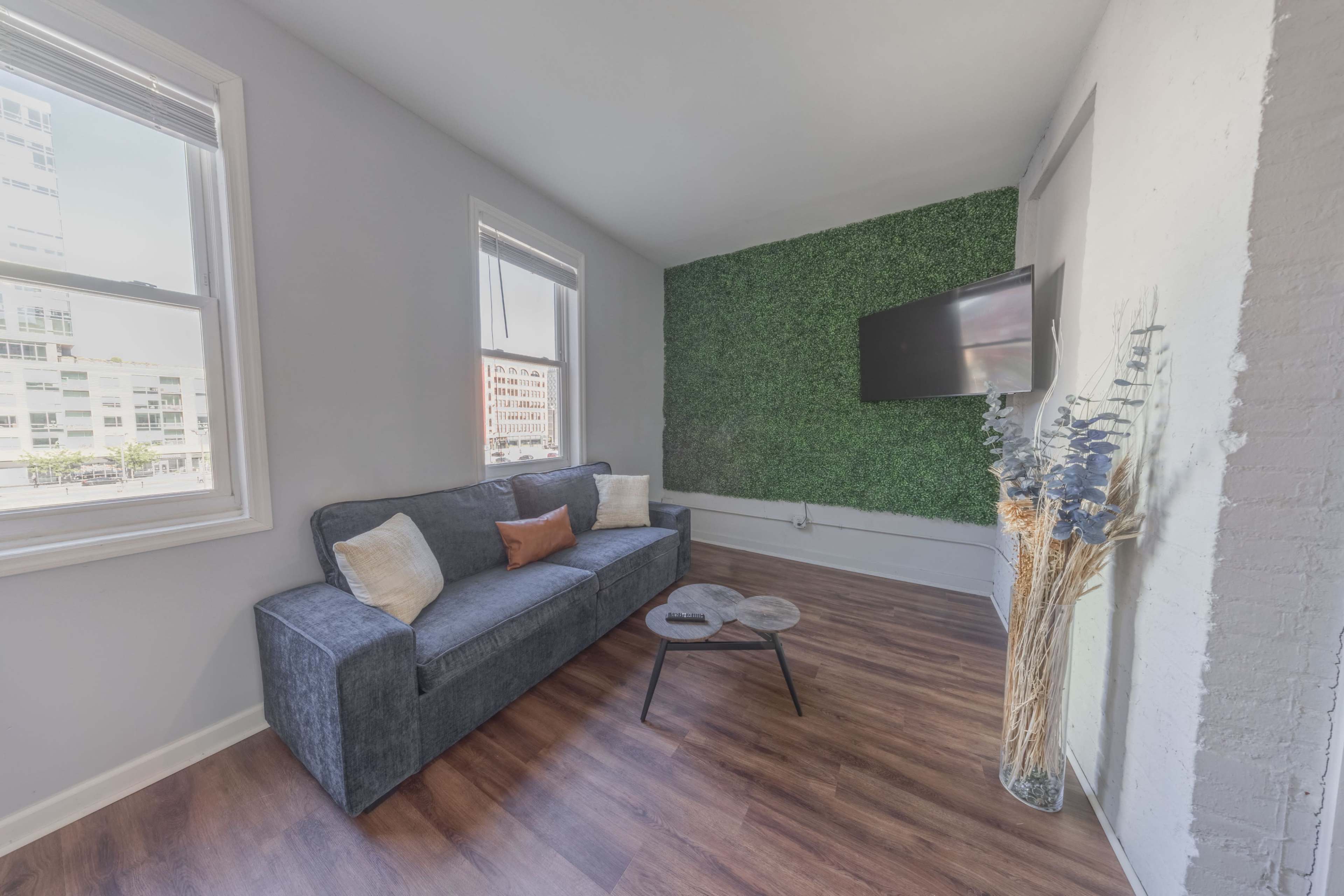 The image shows a modern living room with a gray sofa, a small round coffee table, a television mounted on a green accent wall, and large windows allowing natural light.