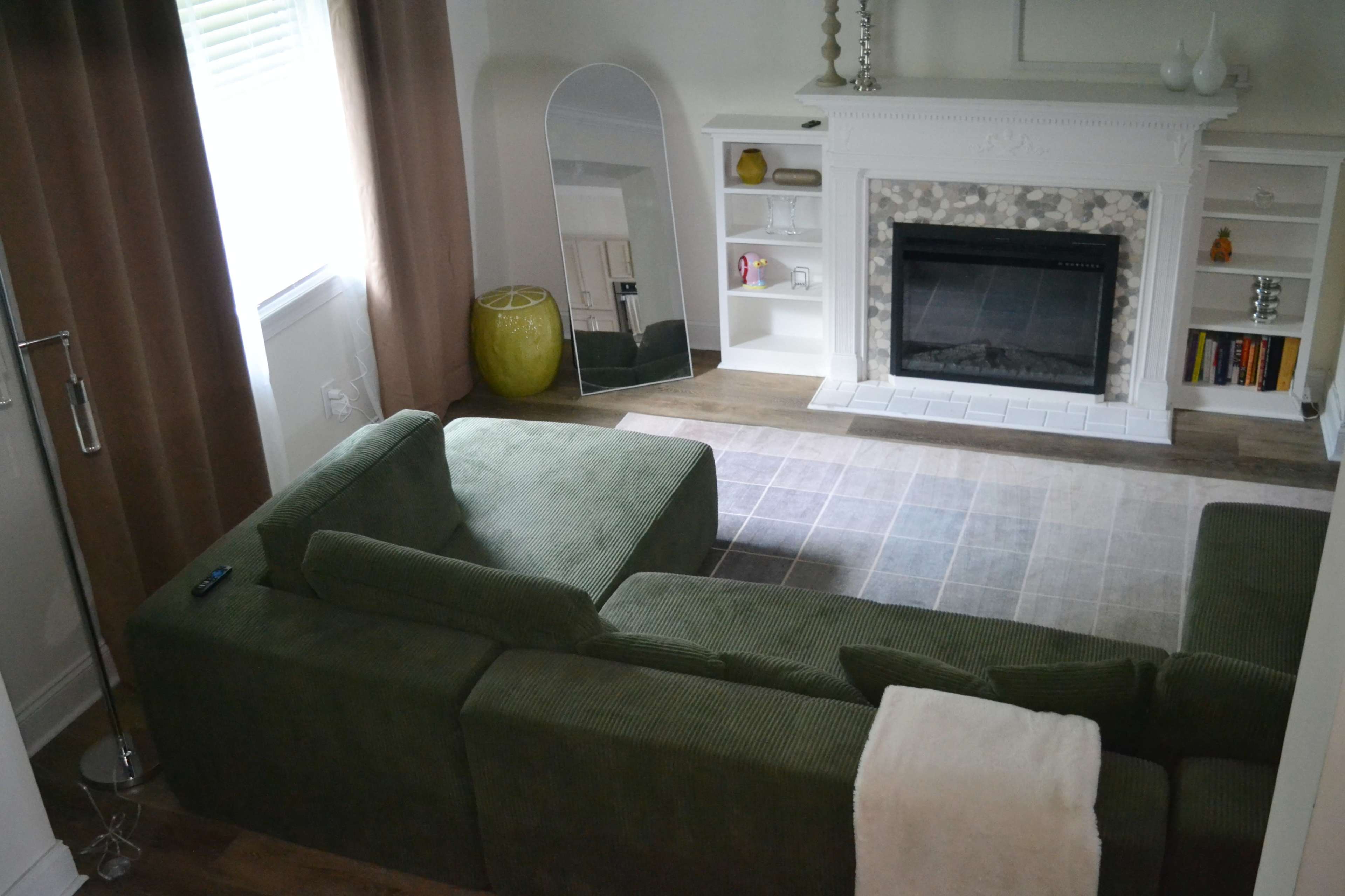 The image shows a cozy living room featuring a green sectional sofa, a mirror, a fireplace with a stone facade, and light-colored furnishings.
