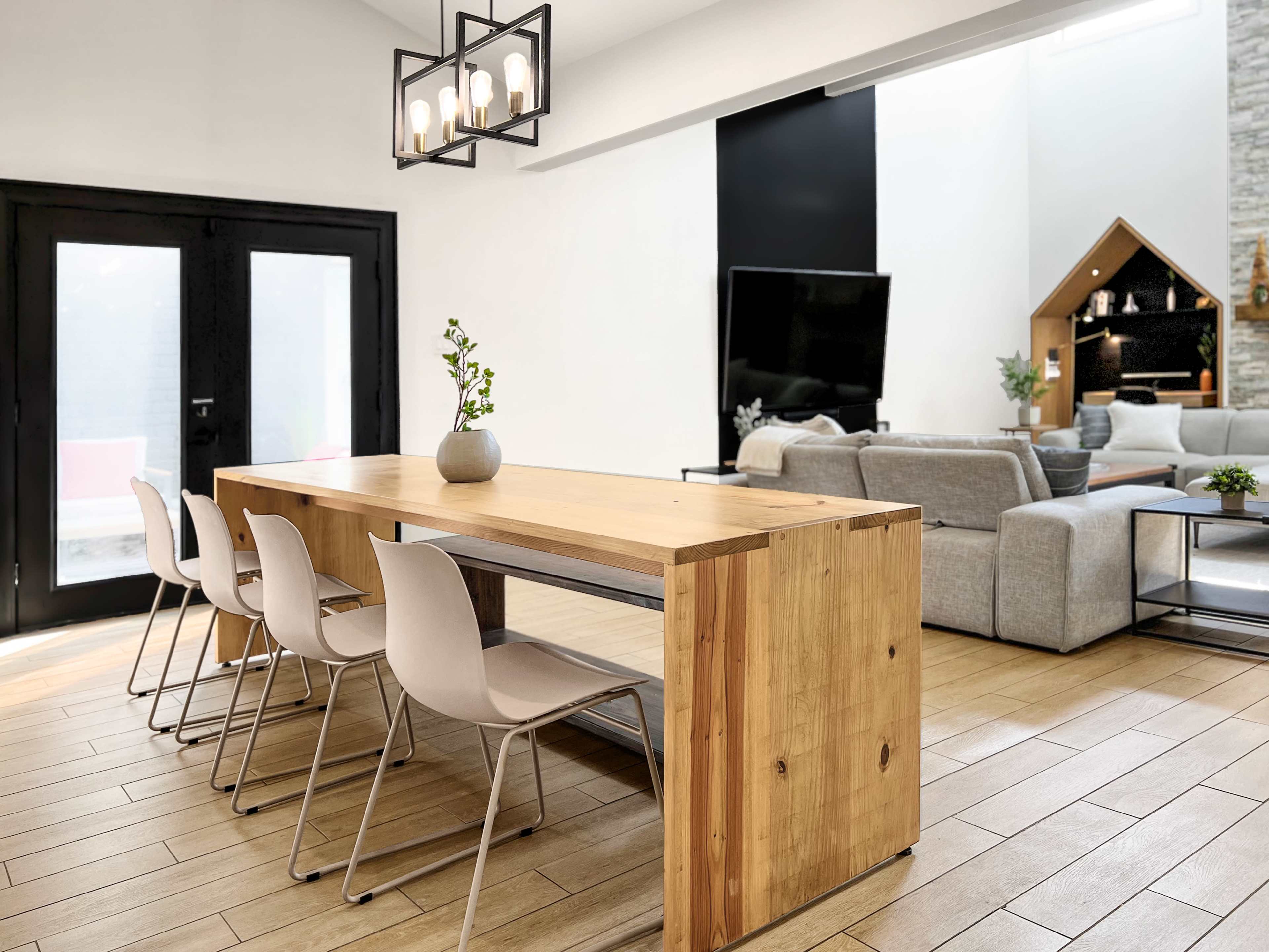 A wooden dining table with four white chairs is positioned in a modern open-concept living area featuring a television and a stone fireplace.