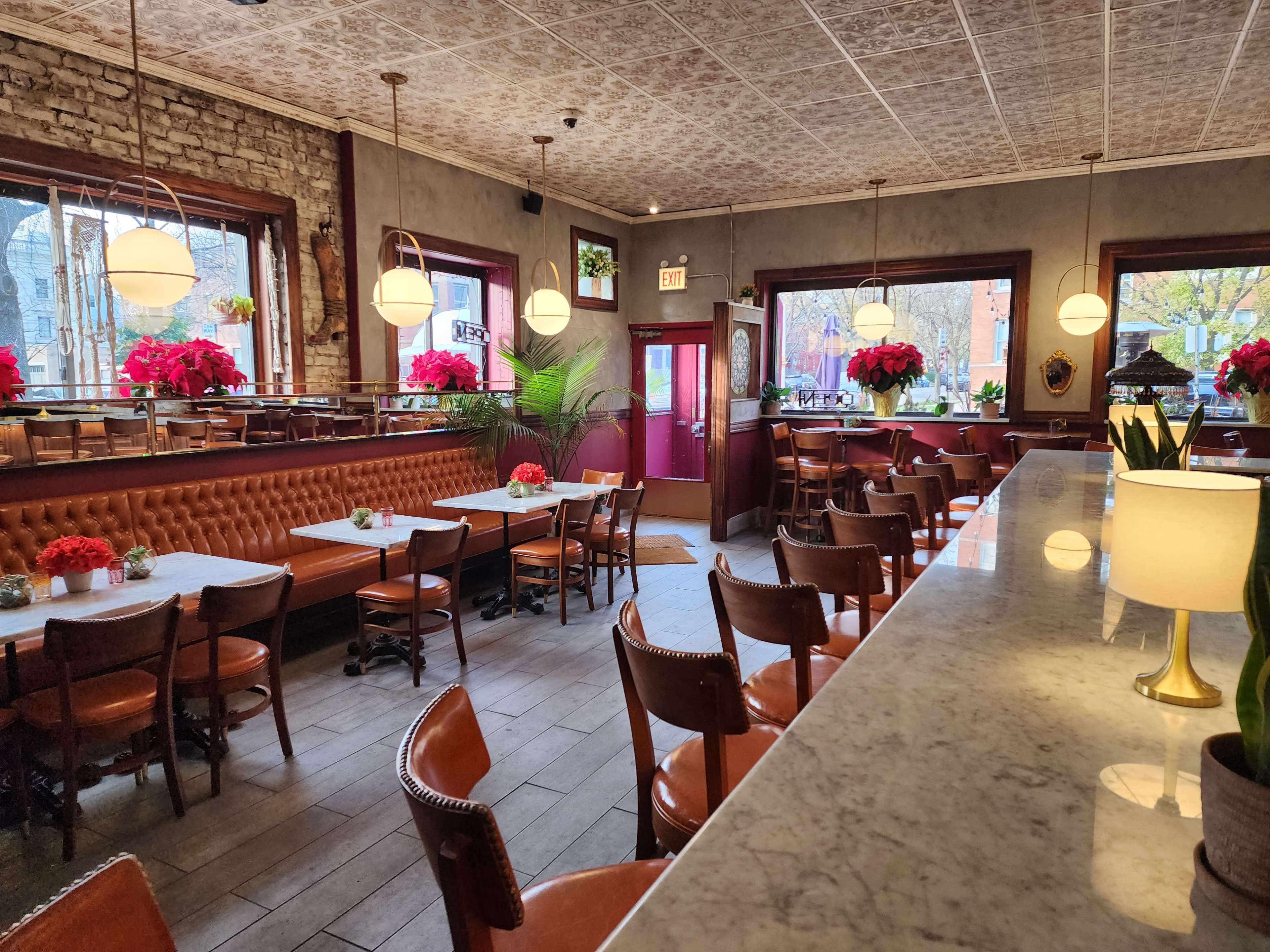 The interior of a restaurant features wooden tables and chairs, a marble countertop, and large windows adorned with red floral arrangements.
