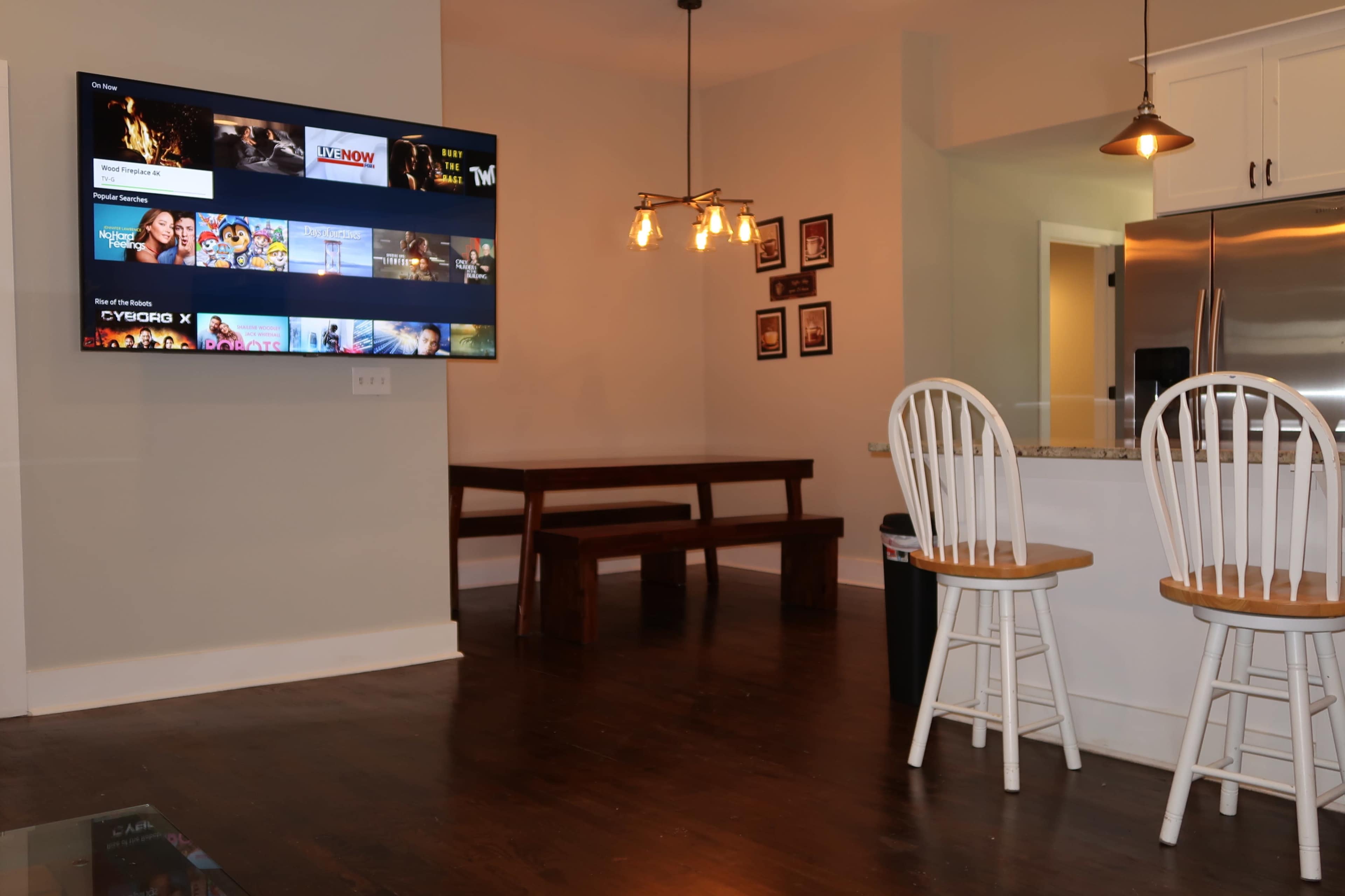 The image shows a modern living space with a wall-mounted television, a wooden dining table with a bench, and two white bar stools positioned by a kitchen counter.