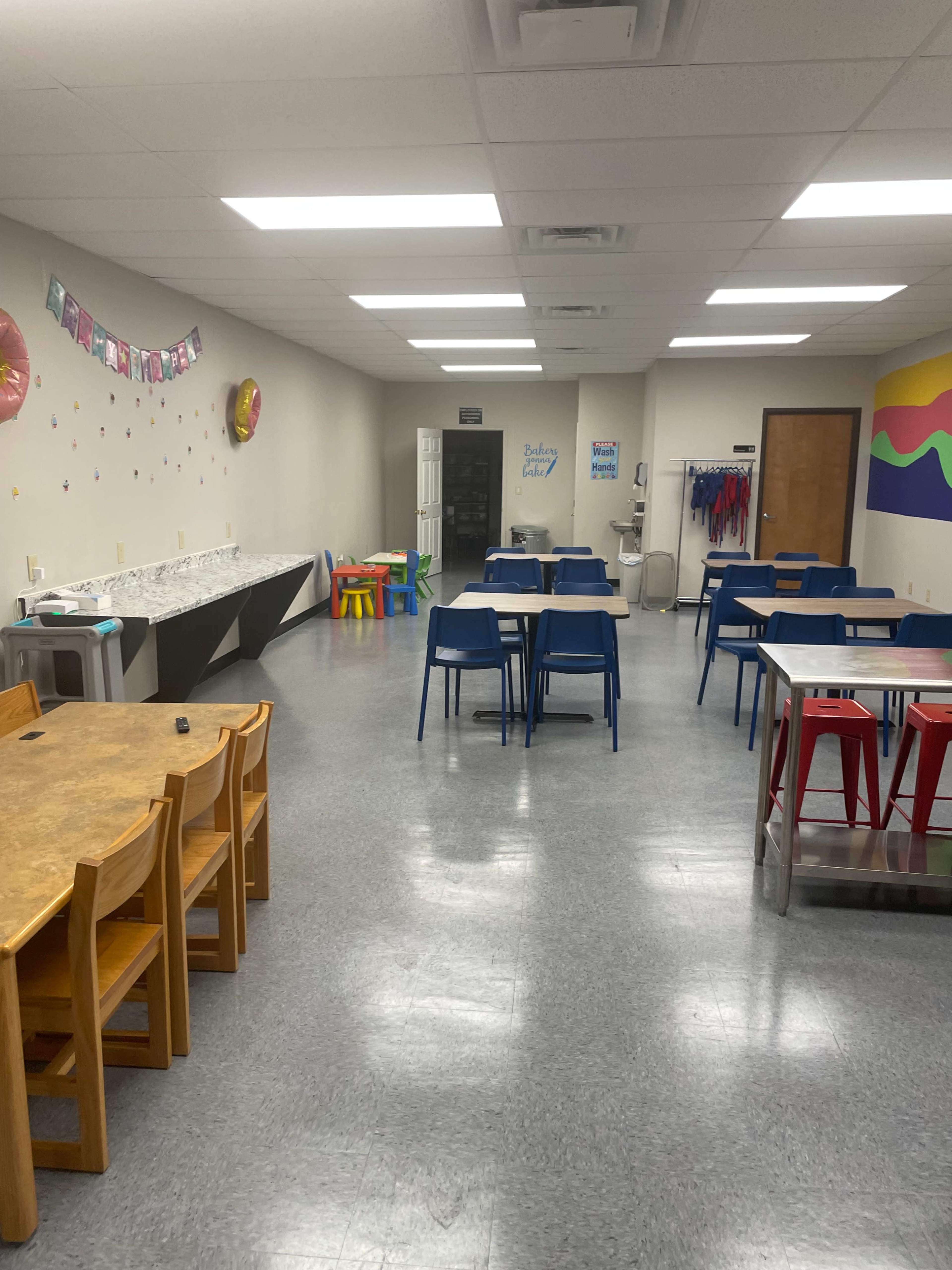 The image shows a spacious classroom with several tables and chairs arranged neatly, along with colorful wall decorations and a water cooler.