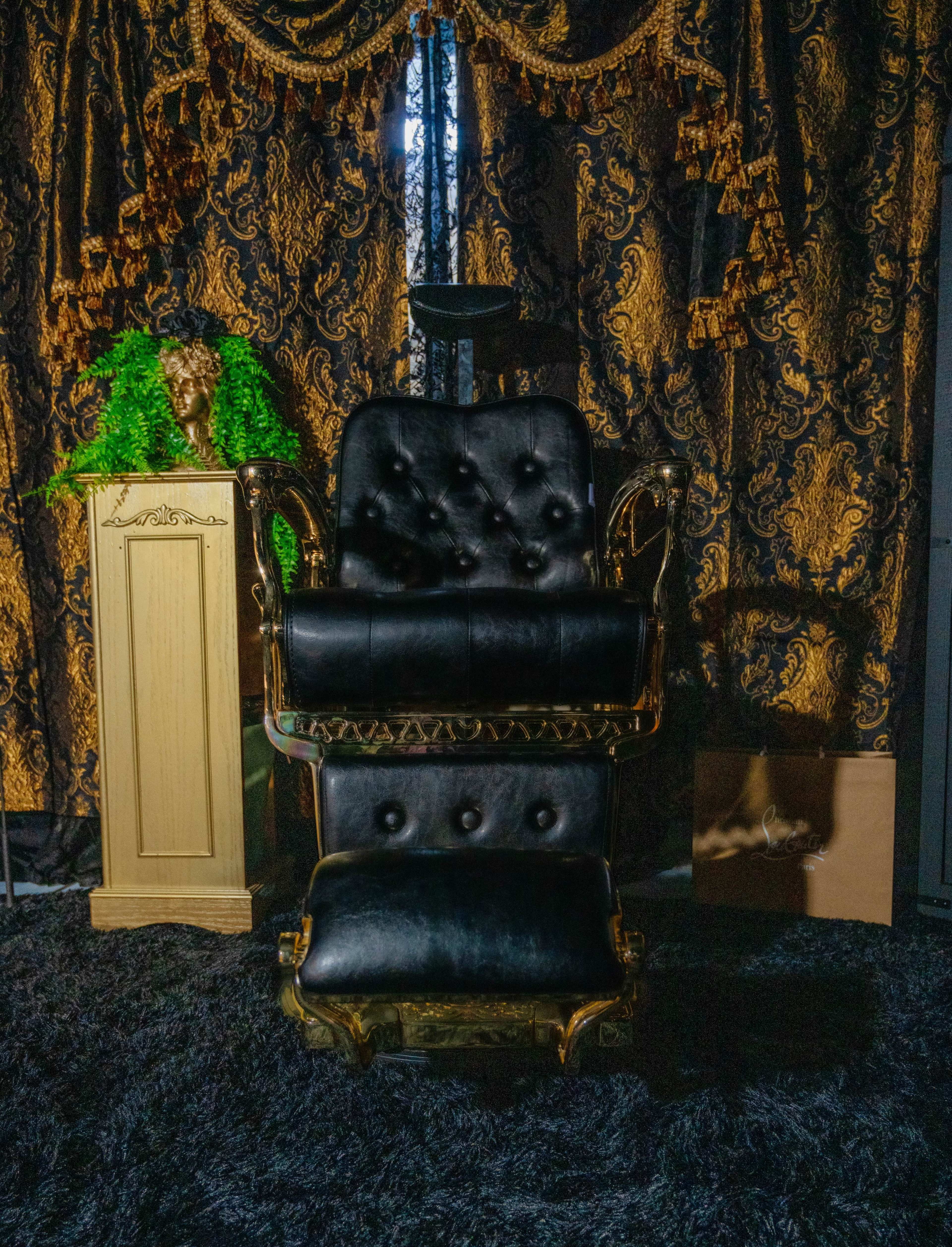 The image features an ornate black leather chair with gold accents positioned in front of dark, patterned curtains, accompanied by a decorative pedestal and a green plant.