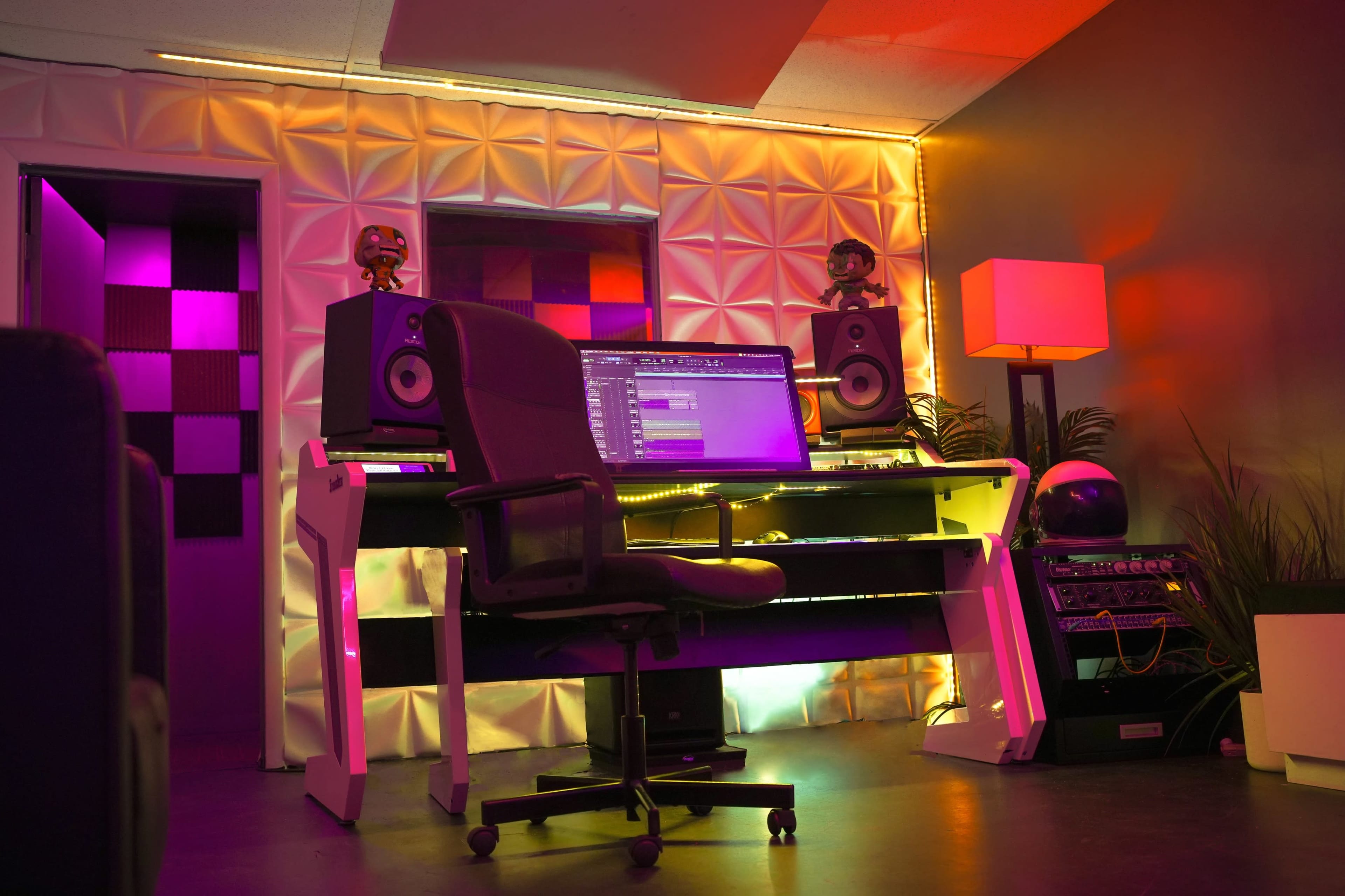 A modern music production studio features a desk with a computer, speakers, a chair, and ambient lighting against a textured wall.