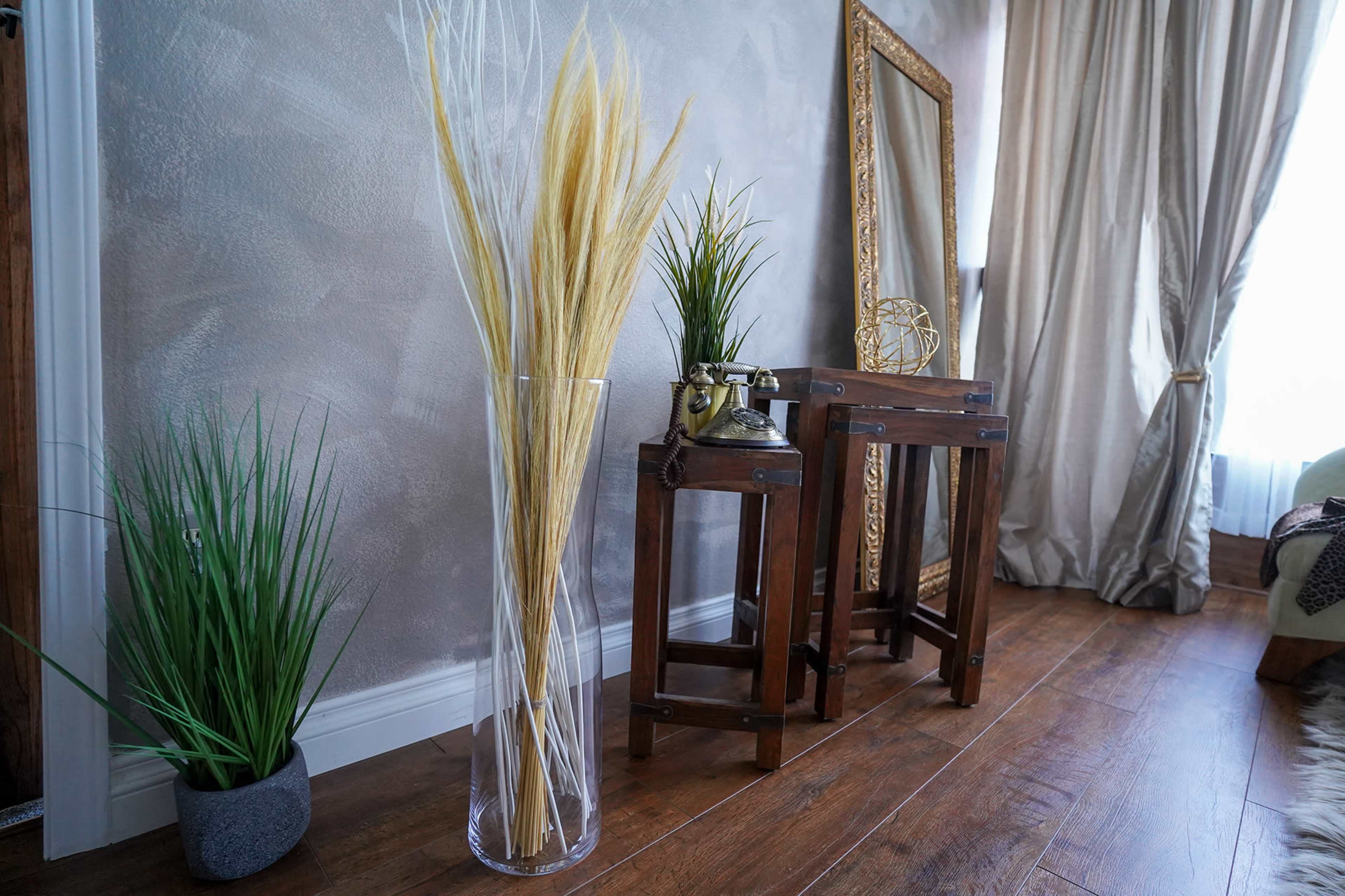 A tall, clear vase with dried grass sits next to a small wooden table and a potted plant against a textured wall, with a large framed mirror and curtains in the background.