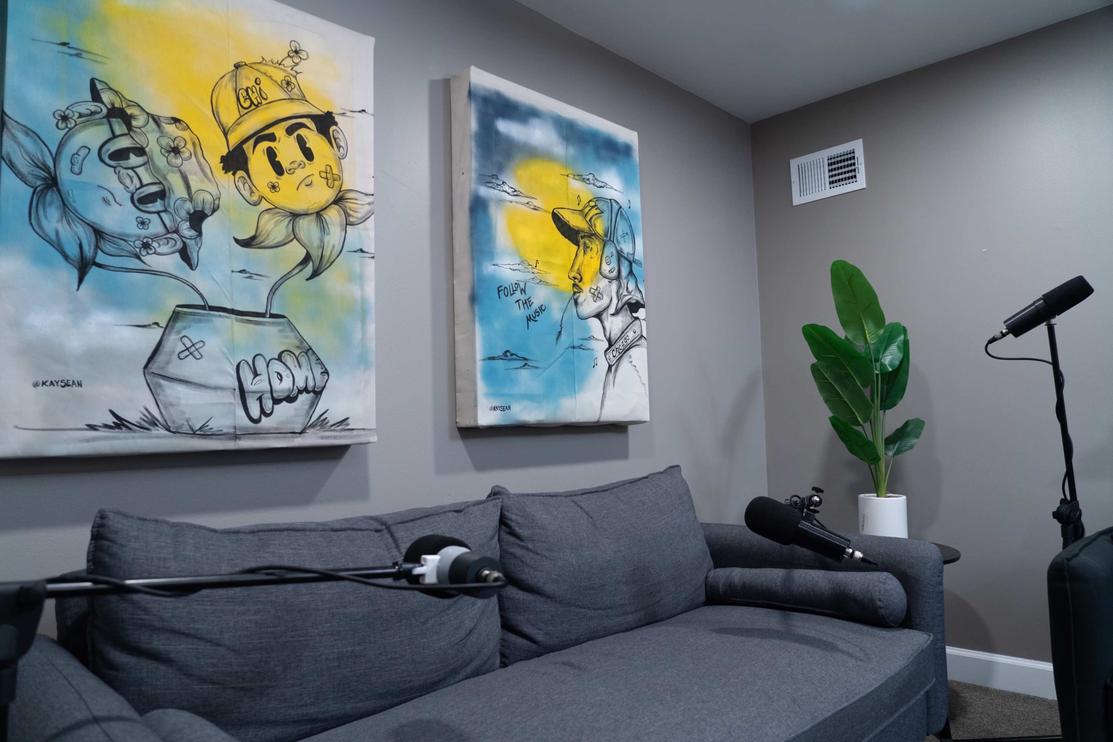 A cozy room featuring a gray couch, two colorful canvas artworks on the wall, a potted plant, and two microphones positioned nearby.