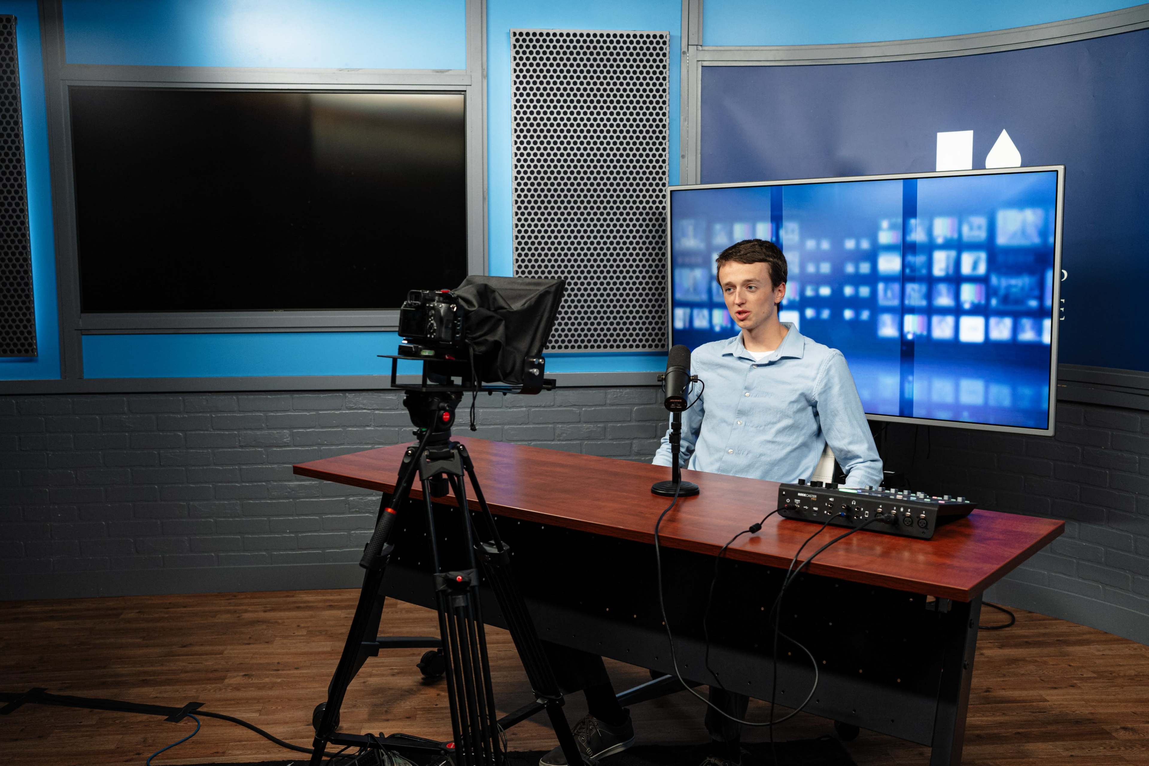 A person sits at a desk in a recording studio, facing a camera and a large television screen displaying multiple images.