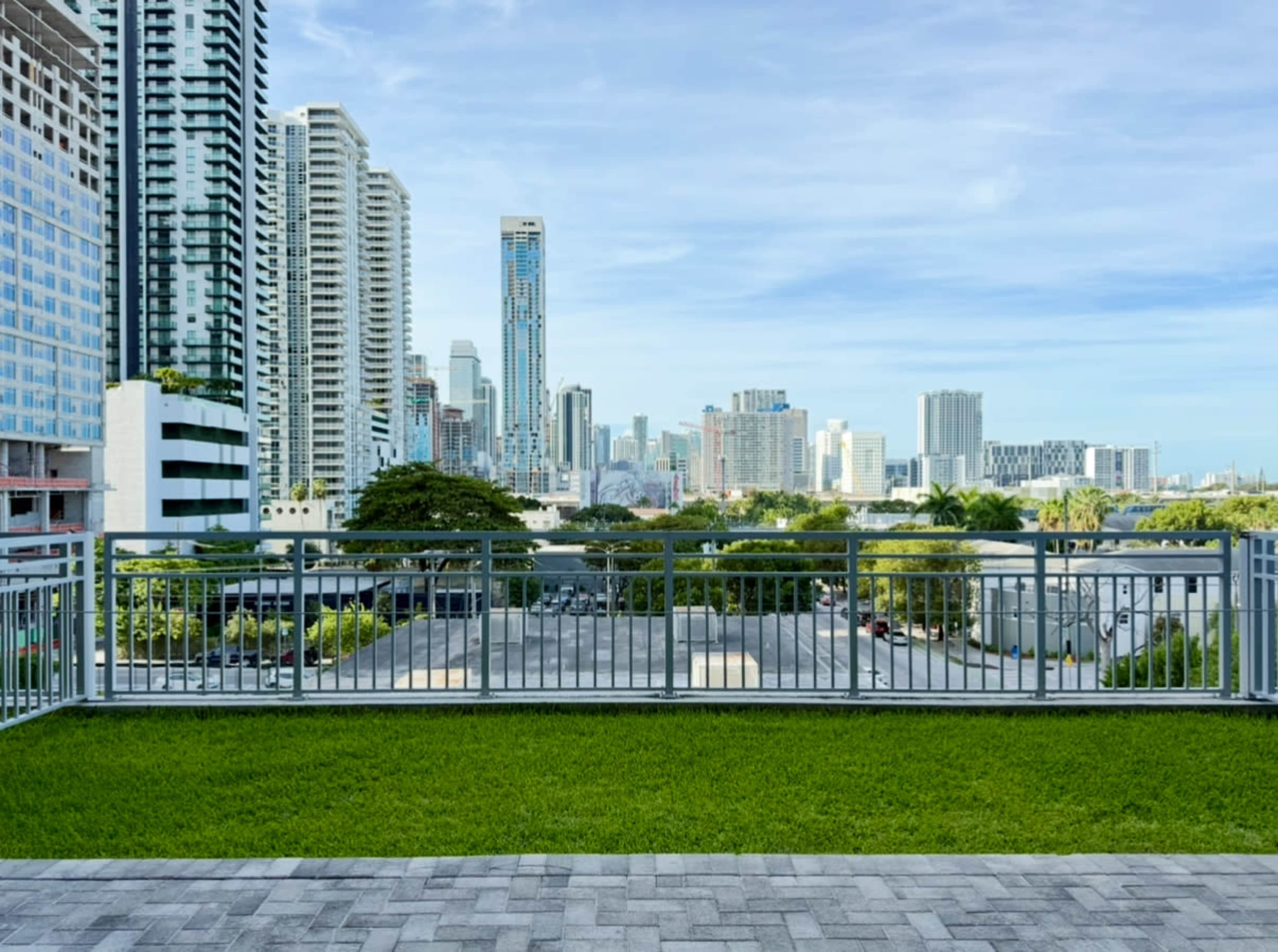 The image shows a modern urban skyline with tall buildings in the background and a green lawn bordered by a metal railing in the foreground.