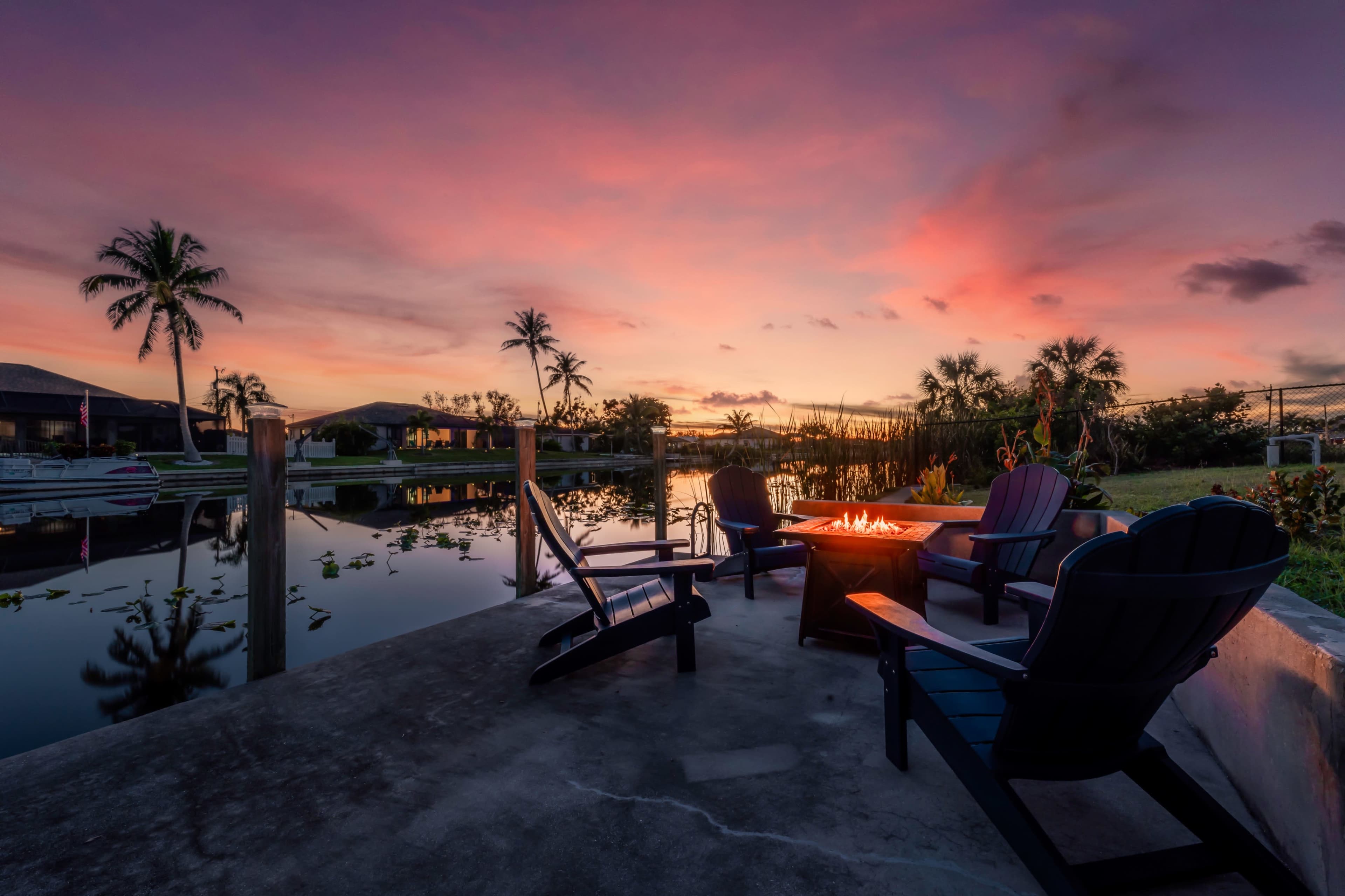 A serene outdoor setting with four Adirondack chairs arranged around a fire pit, overlooking a calm waterway at sunset.