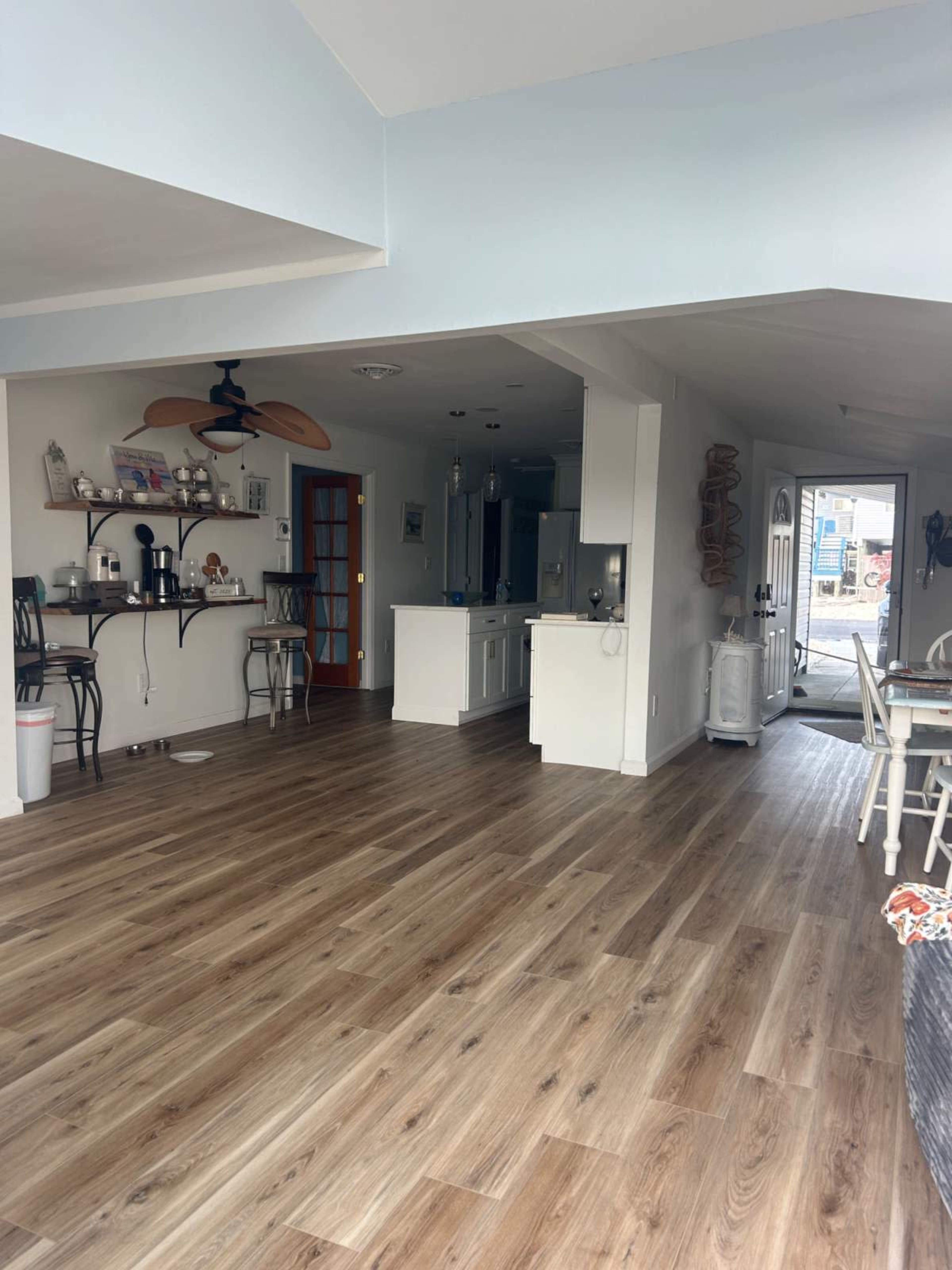 The image shows an open-concept living space featuring a kitchen, dining area, and a doorway leading outside, with wooden flooring and modern furnishings.