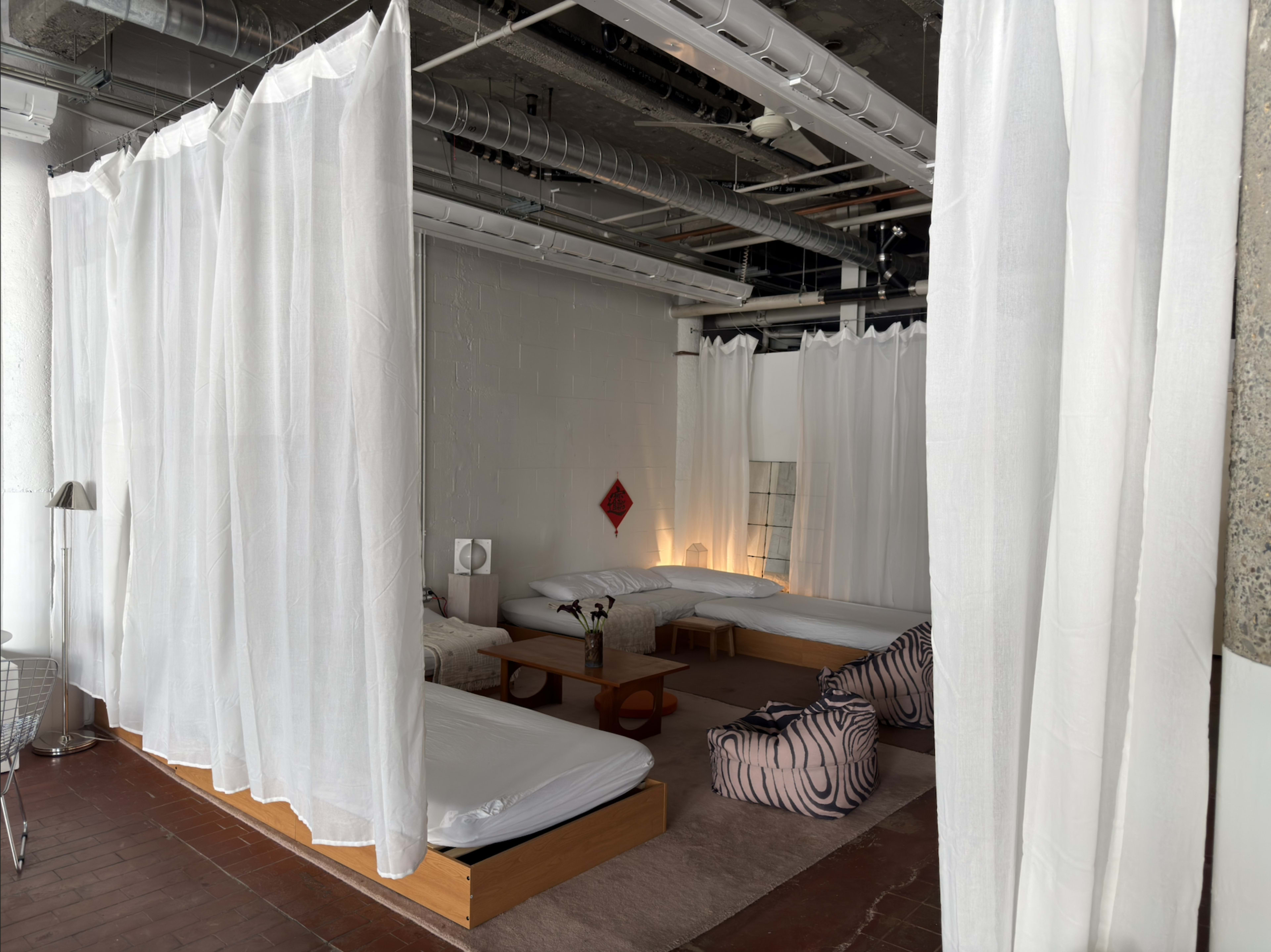The image shows a communal sleeping area with low wooden beds separated by white curtains in an industrial-style room.