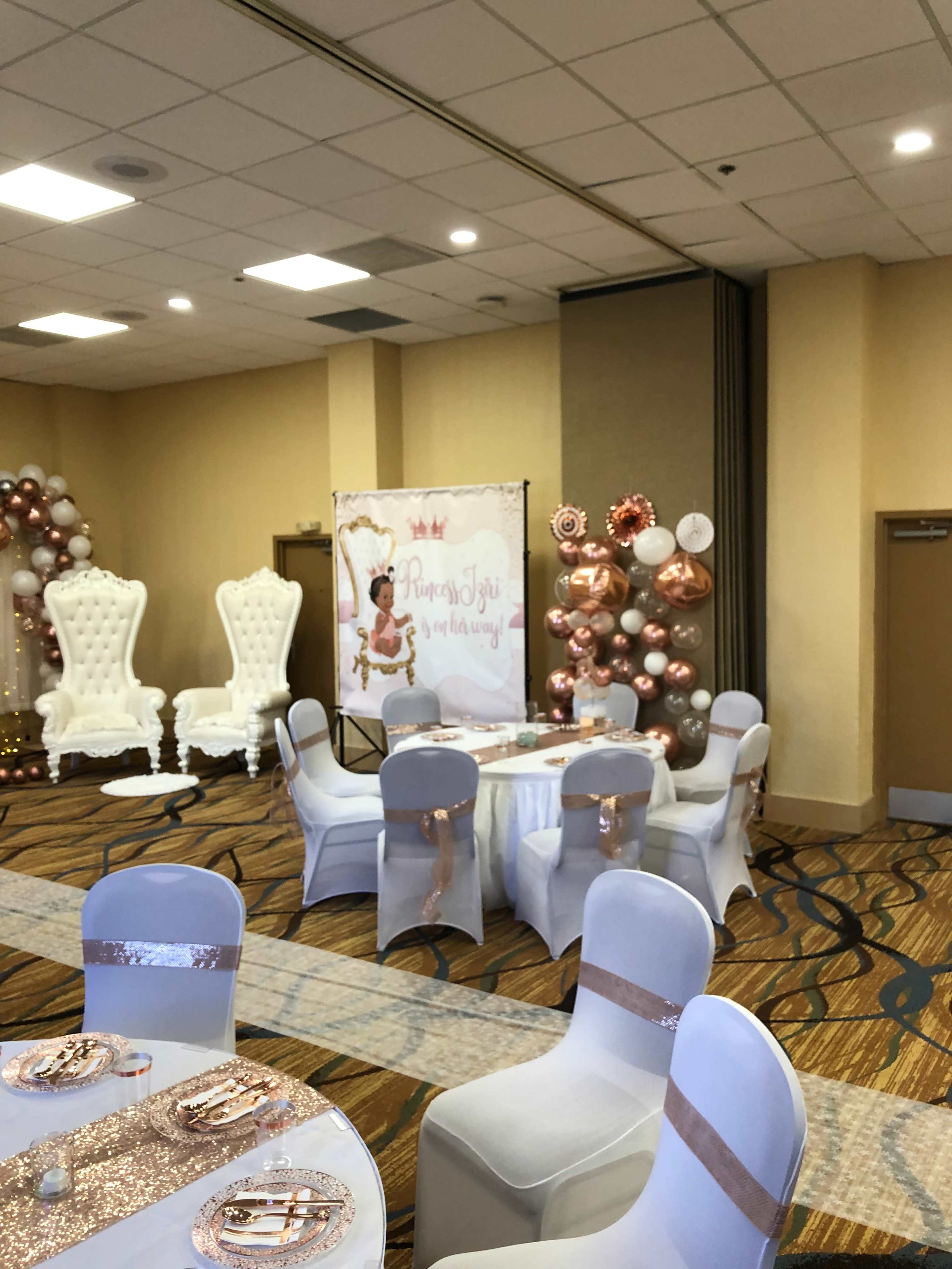 A decorated event space features elegant white chairs, tables set with tableware, and a backdrop with balloons and a themed sign.